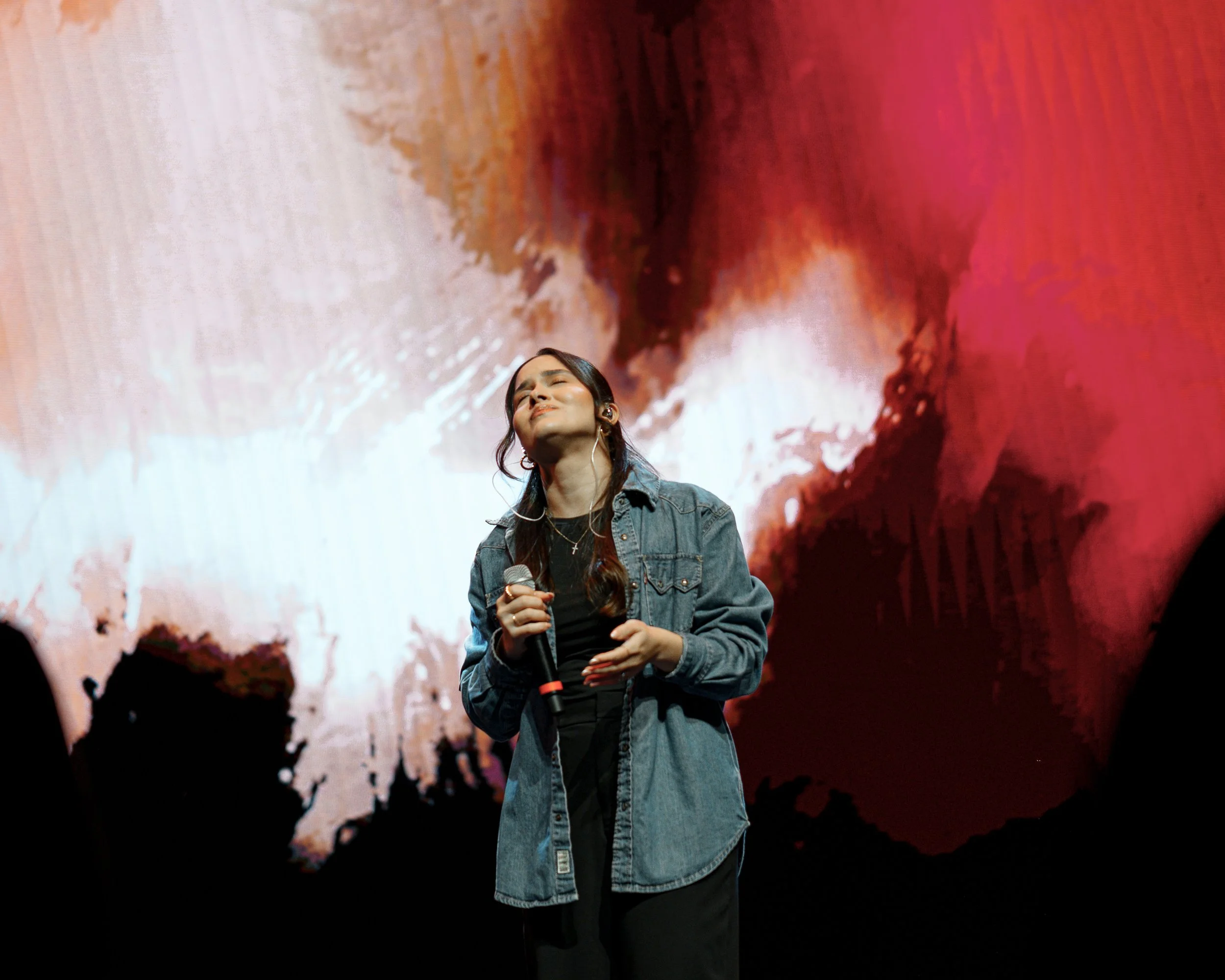 A woman singing on stage holding a microphone with a large colorful abstract background behind her.