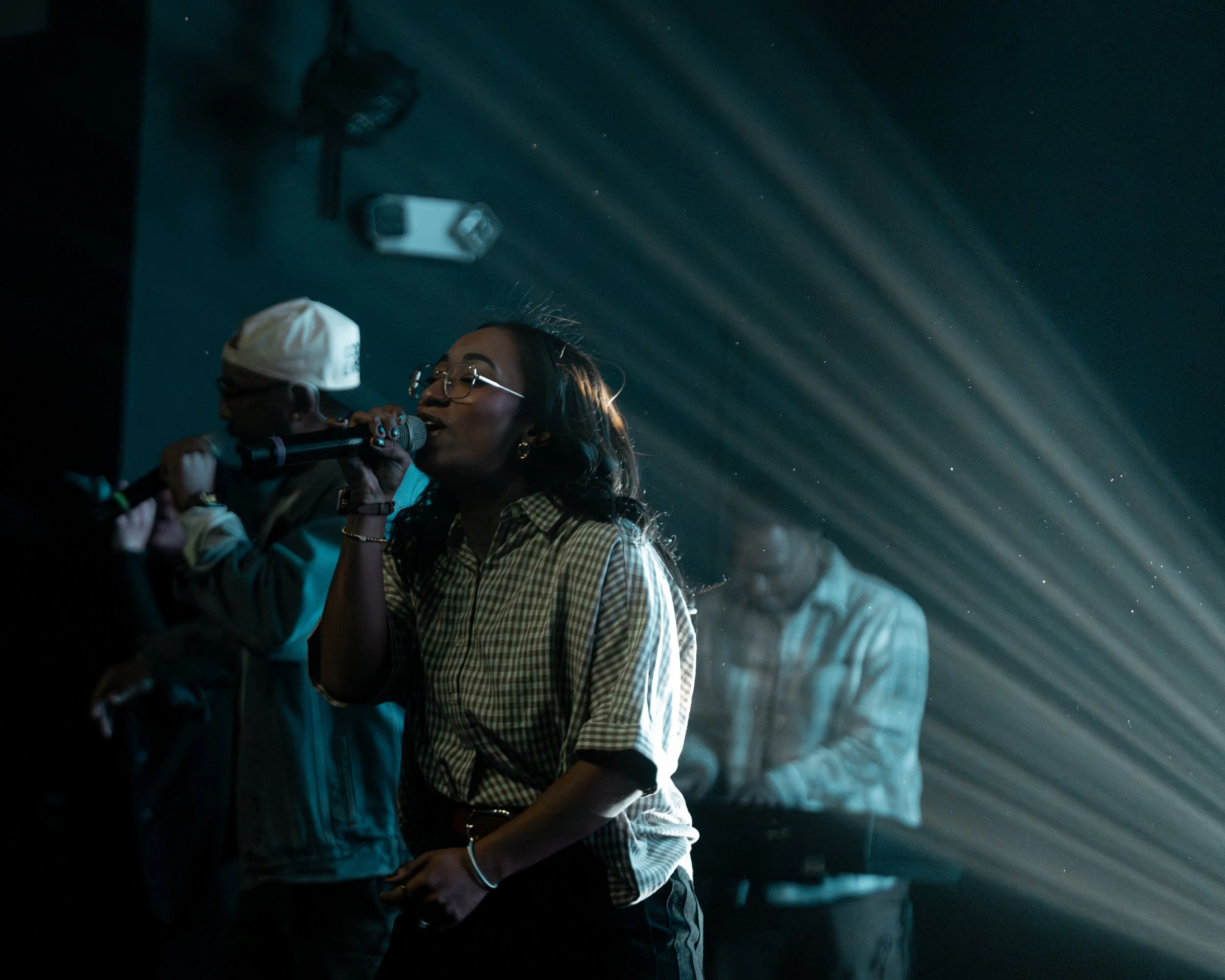 A woman with glasses singing into a microphone, with a man in a white hat and a person at a keyboard visible in the background. Stage lighting creates a beams of light effect.