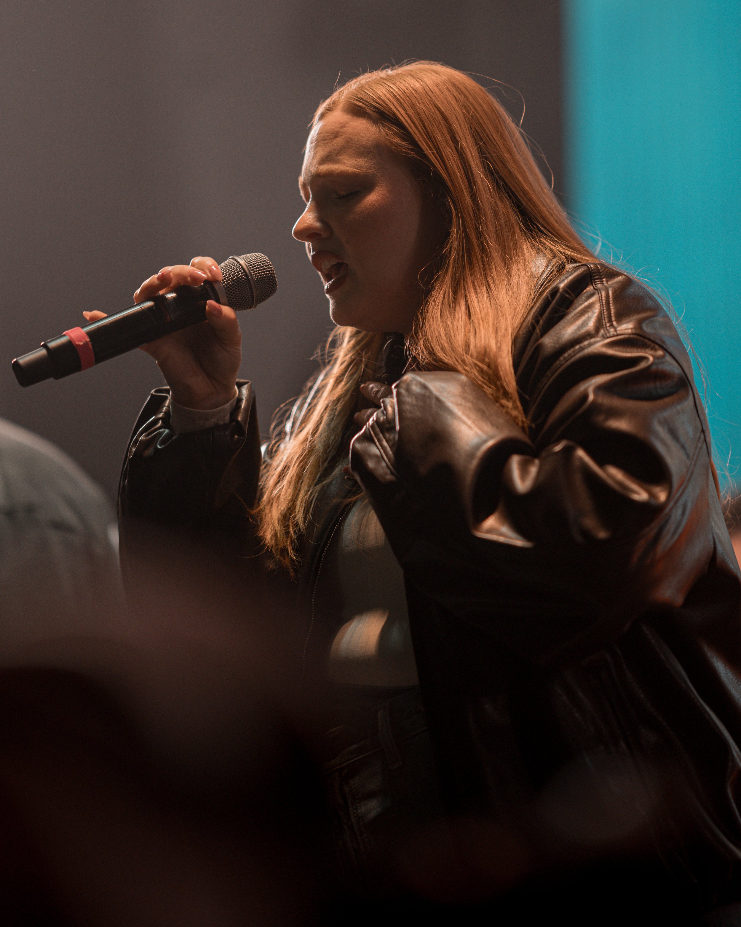 A woman with long red hair singing into a microphone, wearing a black leather jacket, on stage.