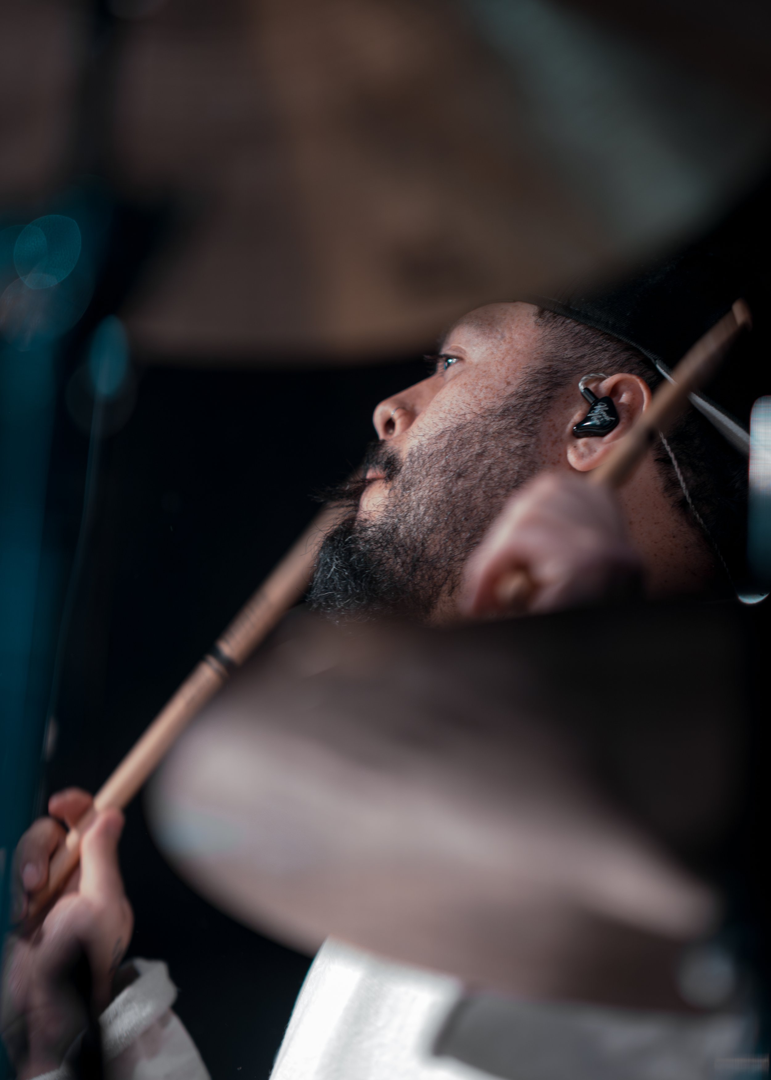 Close-up of a drummer playing, wearing a black cap and an in-ear monitor, with a brown drumstick in hand, captured in side profile.