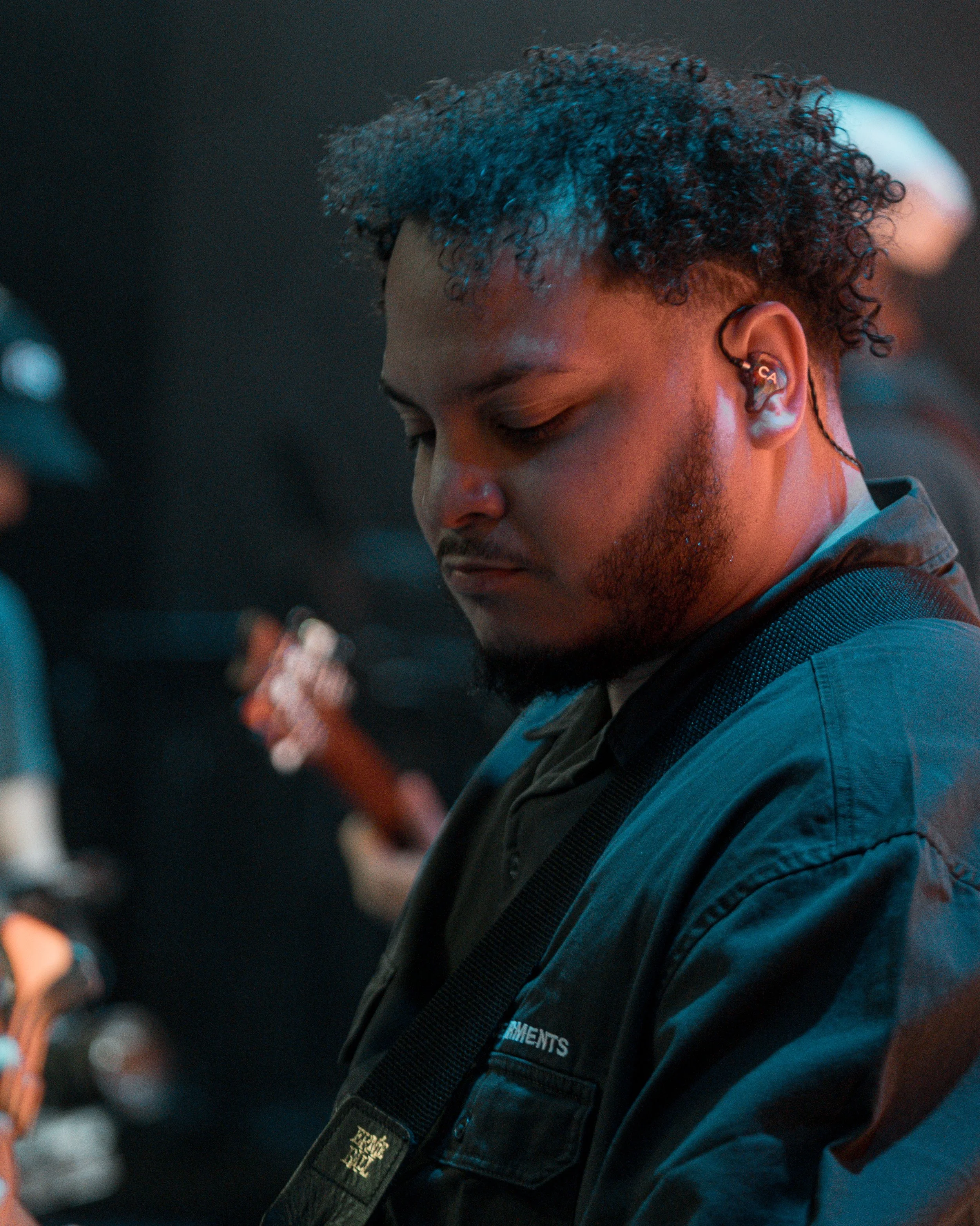 A man with curly hair and a beard looking down, wearing an in-ear monitor and a dark jacket, in a dimly lit setting possibly backstage or at a concert.