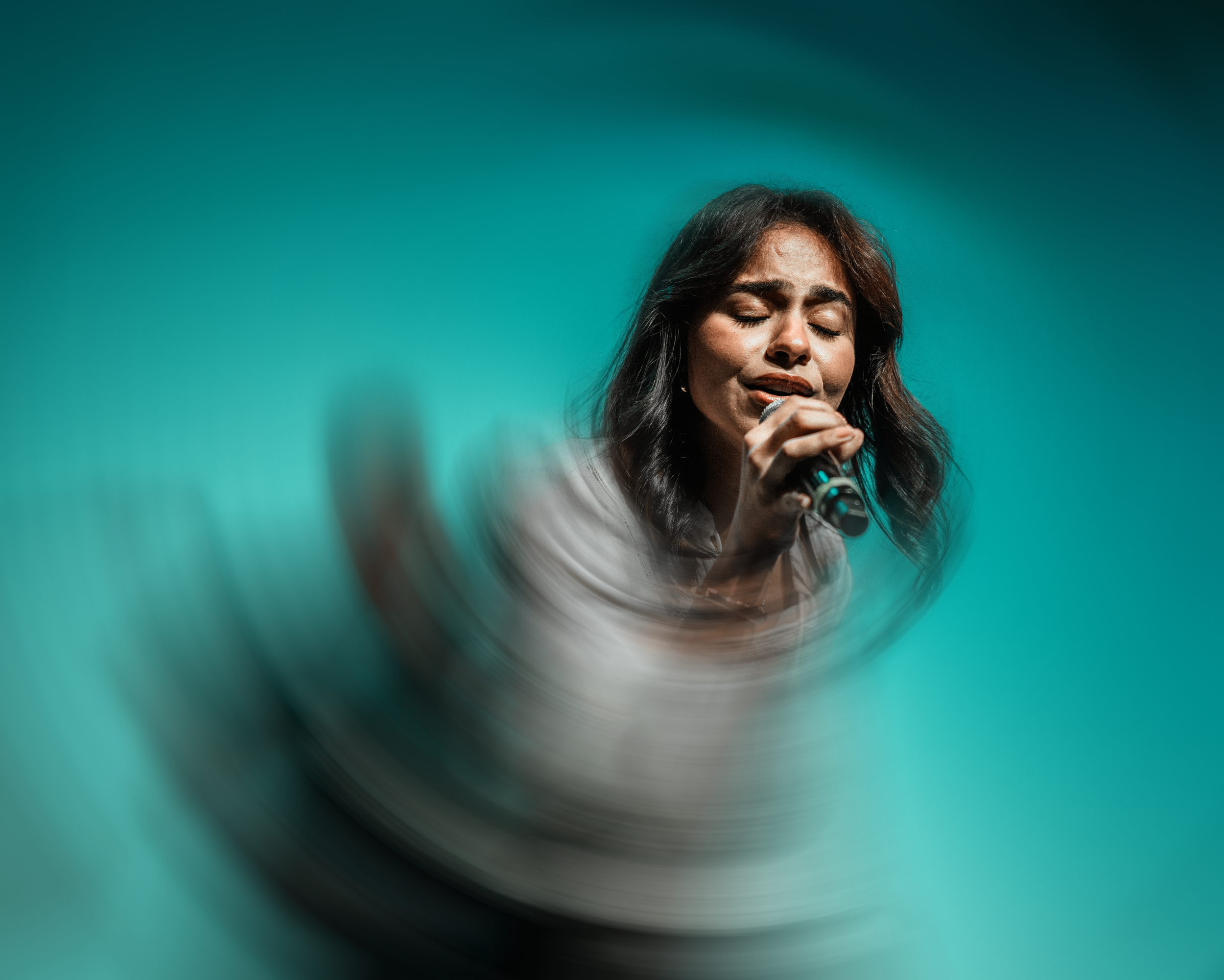 A woman singing passionately with her eyes closed, holding a microphone, with a motion blur effect around her against a turquoise background.