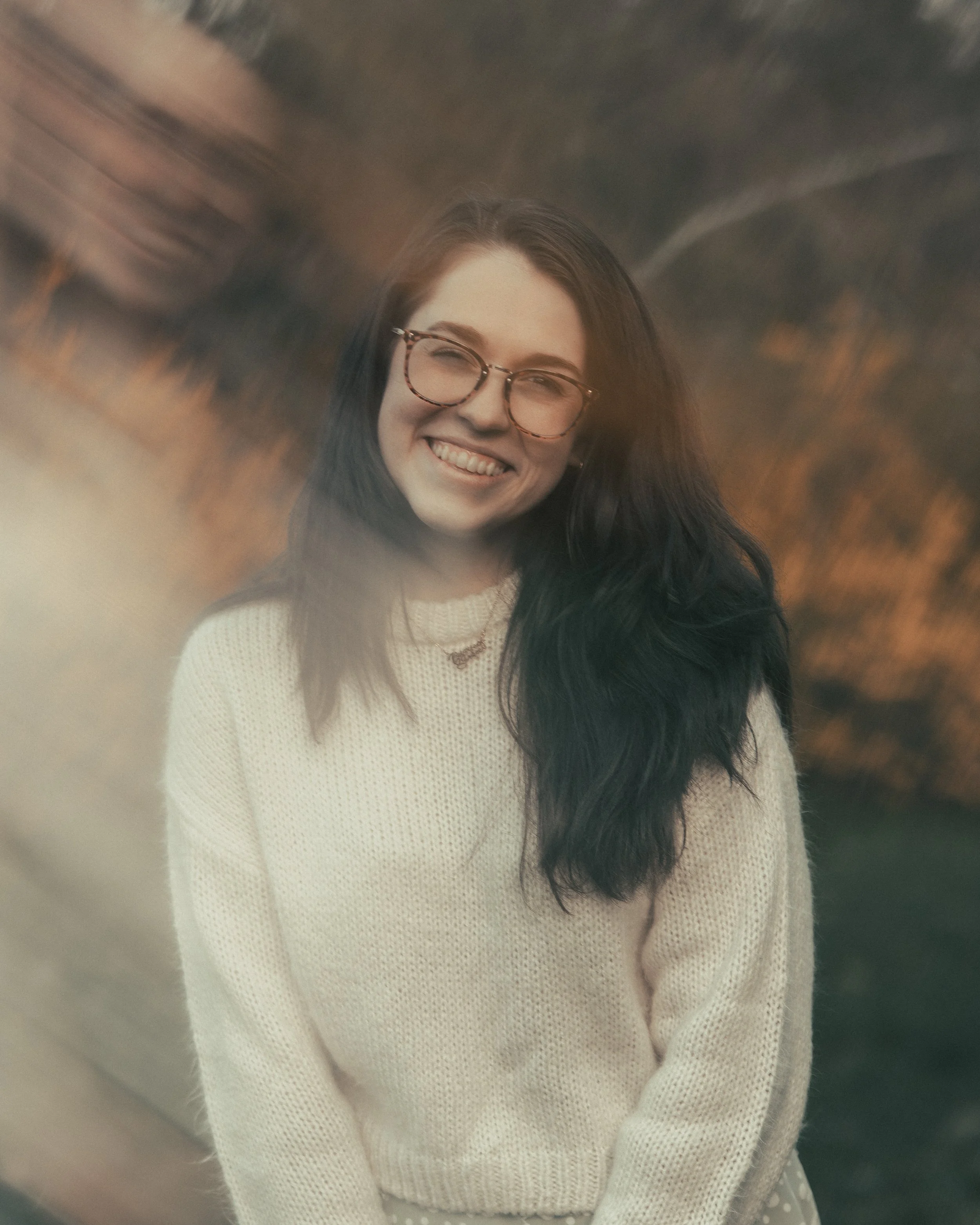 A young woman with glasses smiling outdoors during sunset, wearing a cream sweater and a necklace.