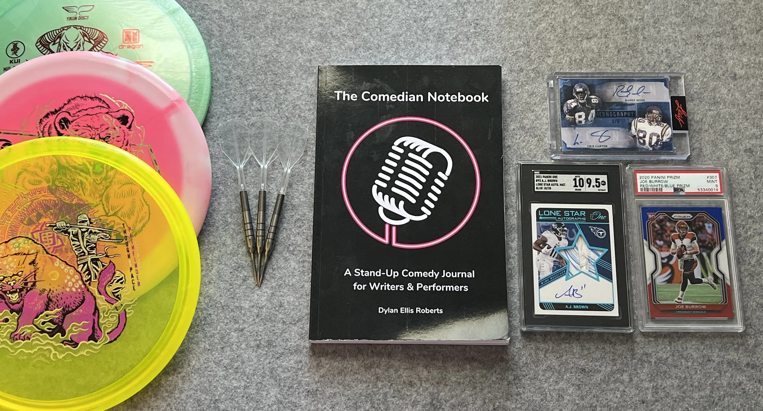 A collection of discs, darts, a journal titled 'The Comedian Notebook,' and sports cards displayed - My hobbies!