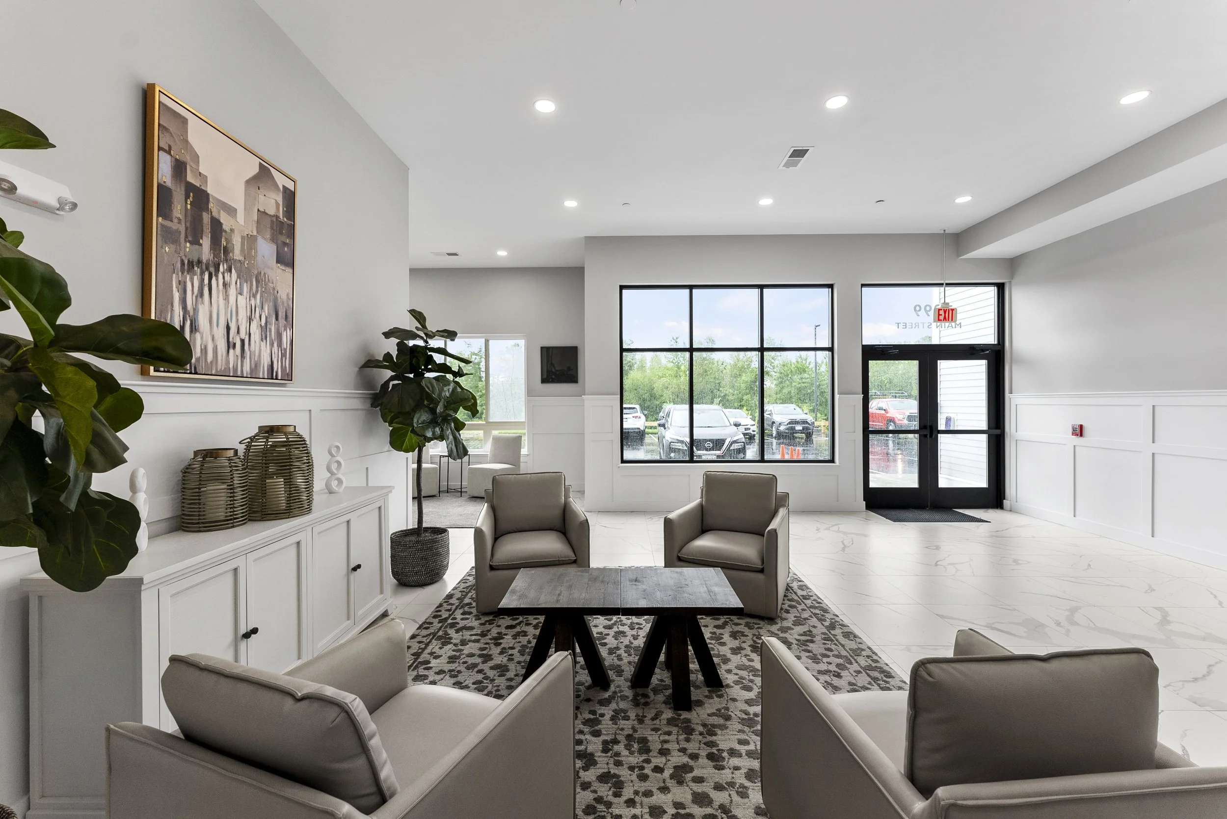 Modern waiting room with beige chairs, a dark wooden table, large window, parking lot view, white walls, art painting, indoor plants, and entry door.
