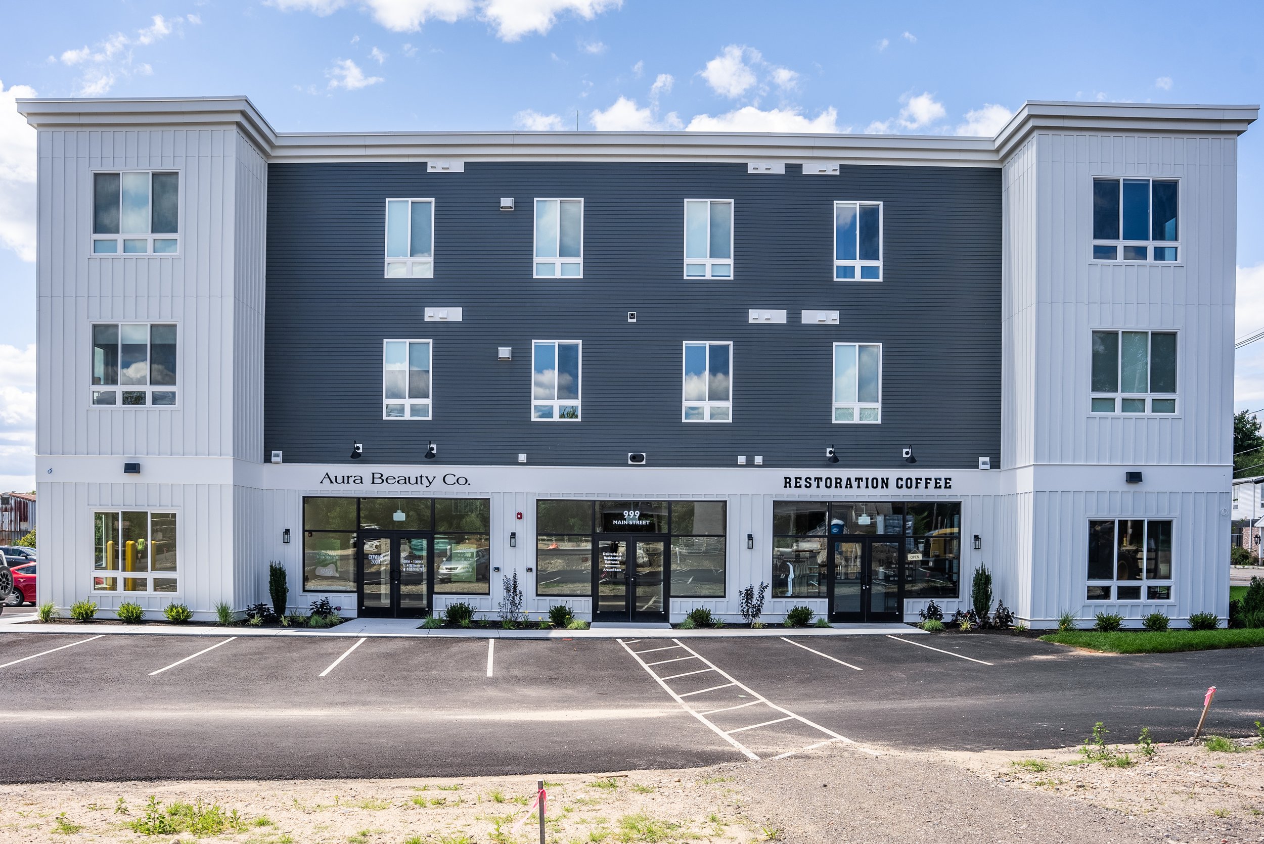 New three-story commercial building with white and dark gray exterior, large front windows, a parking lot with marked spaces, and a blue sky with clouds.