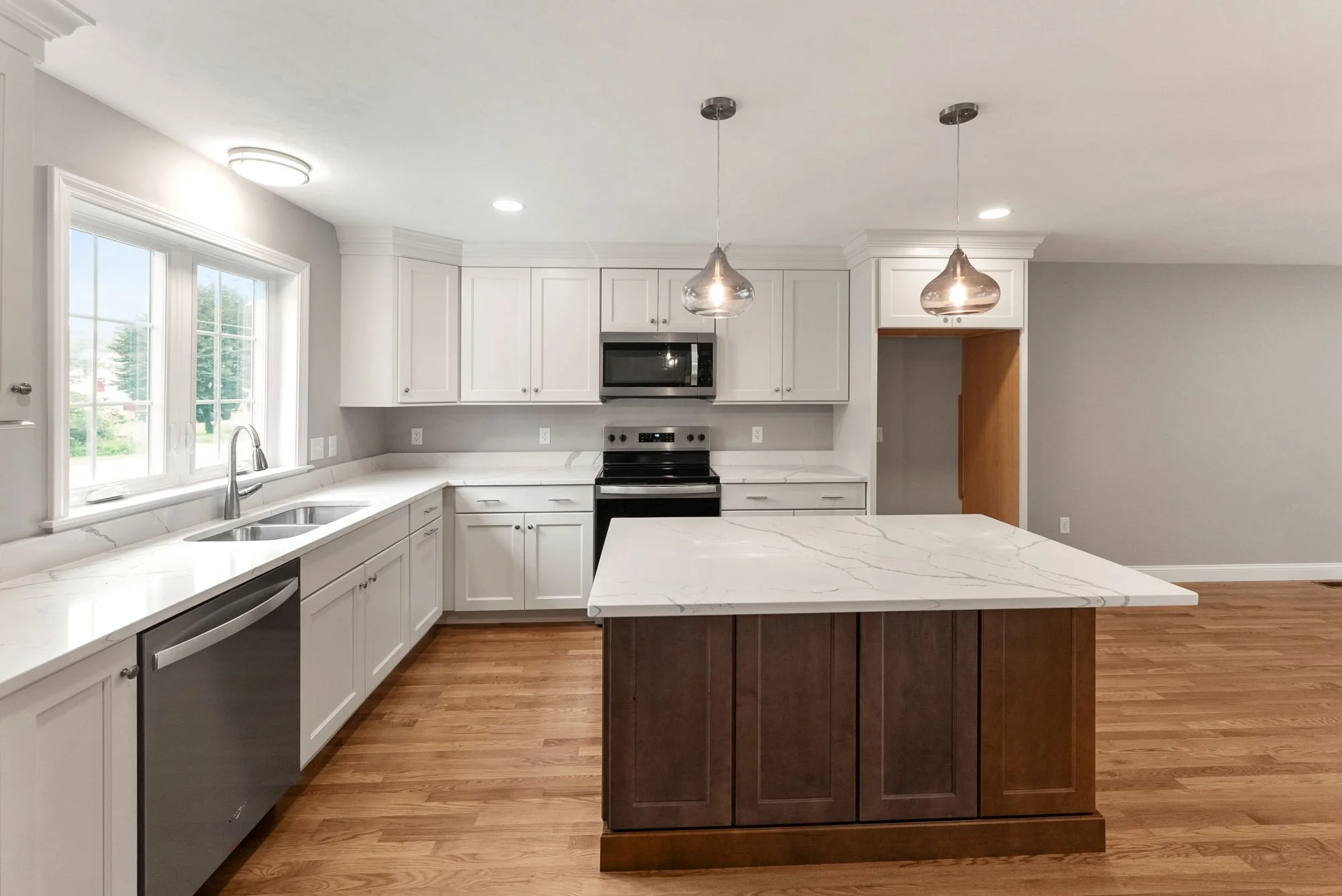 Modern white kitchen with wood flooring, large window, central island with marble countertop, stainless steel appliances, and hanging pendant lights.