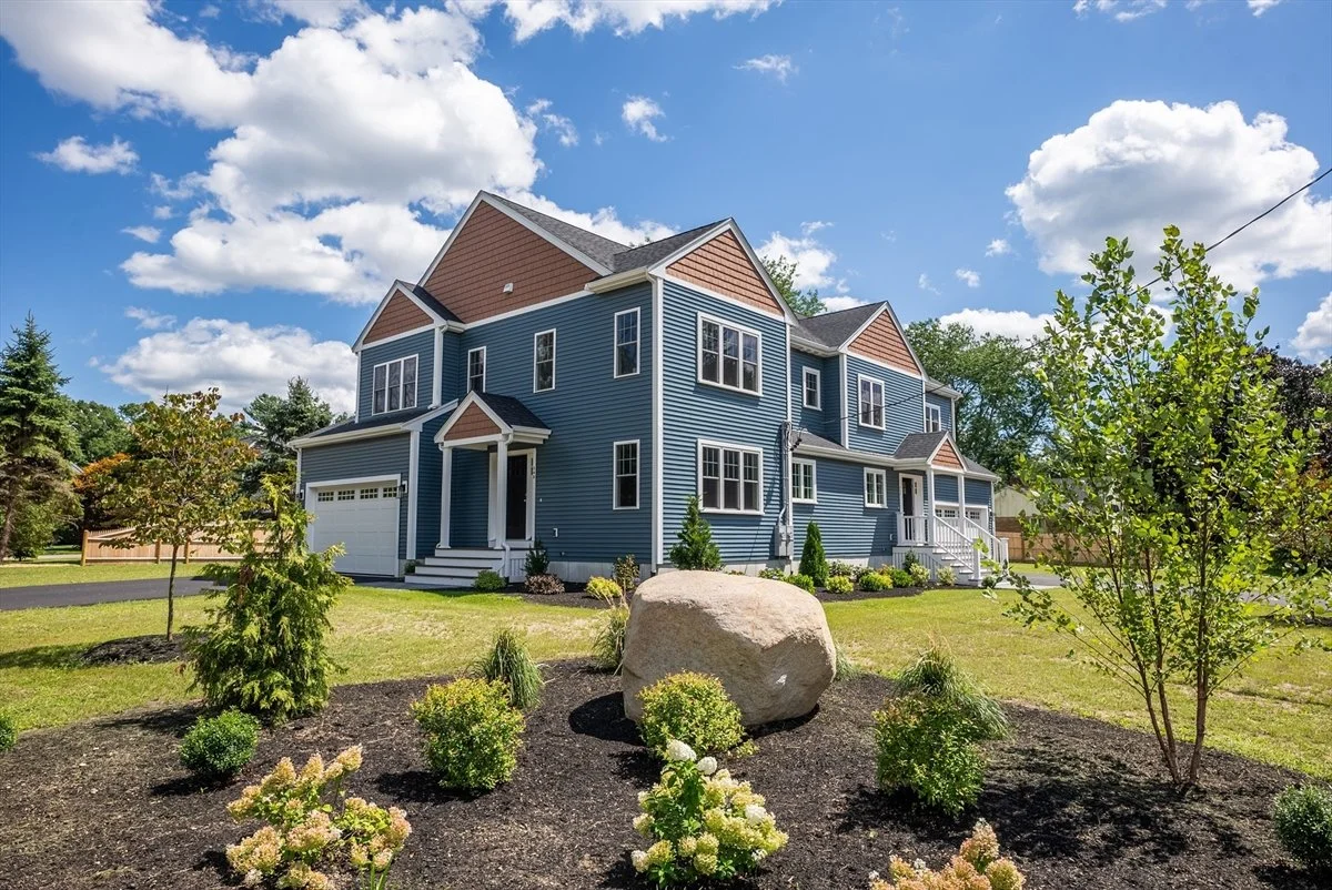 A large, modern blue house with white trim, surrounded by a landscaped yard with small bushes and trees, under a partly cloudy sky.