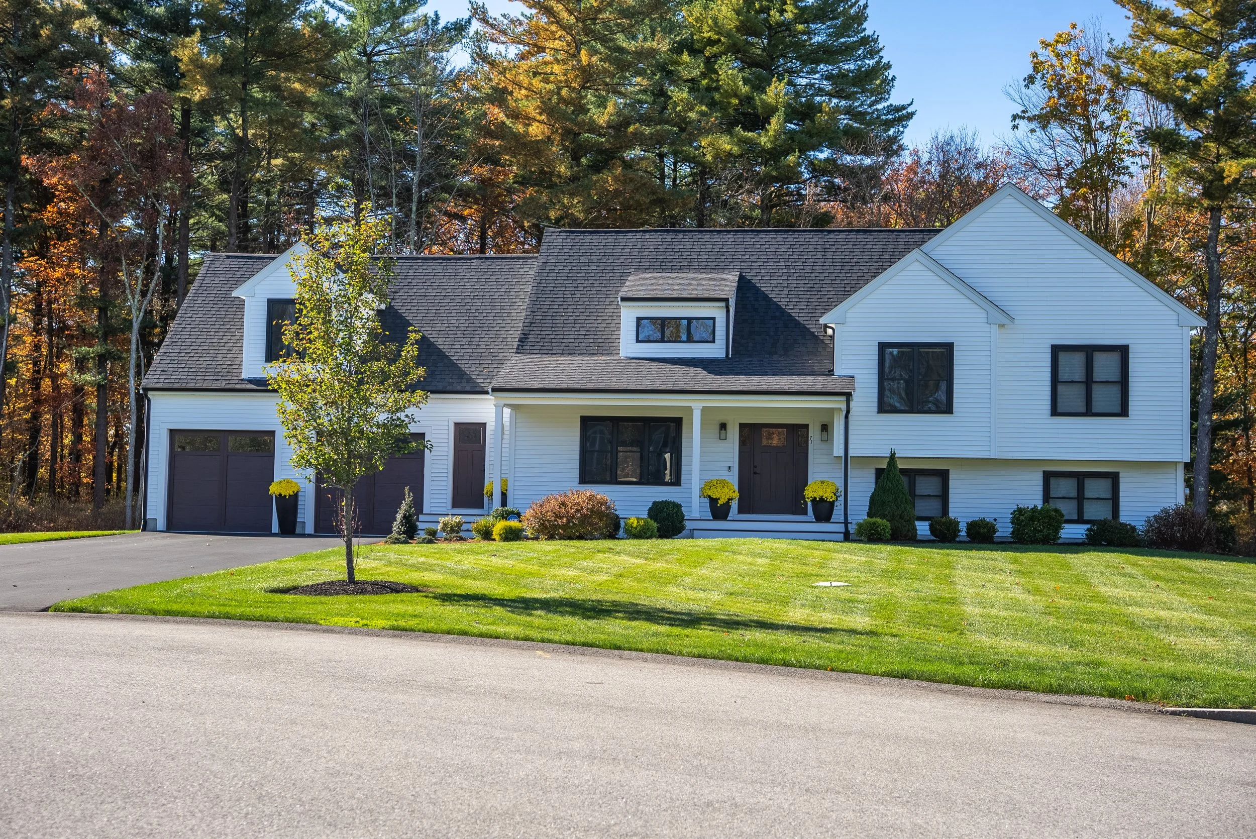 A two-story house with white siding, black window frames, and a dark shingled roof. The front yard has a small tree, neatly trimmed lawn, and potted plants with yellow flowers near the entrance. There is a paved driveway leading to a garage on the left.