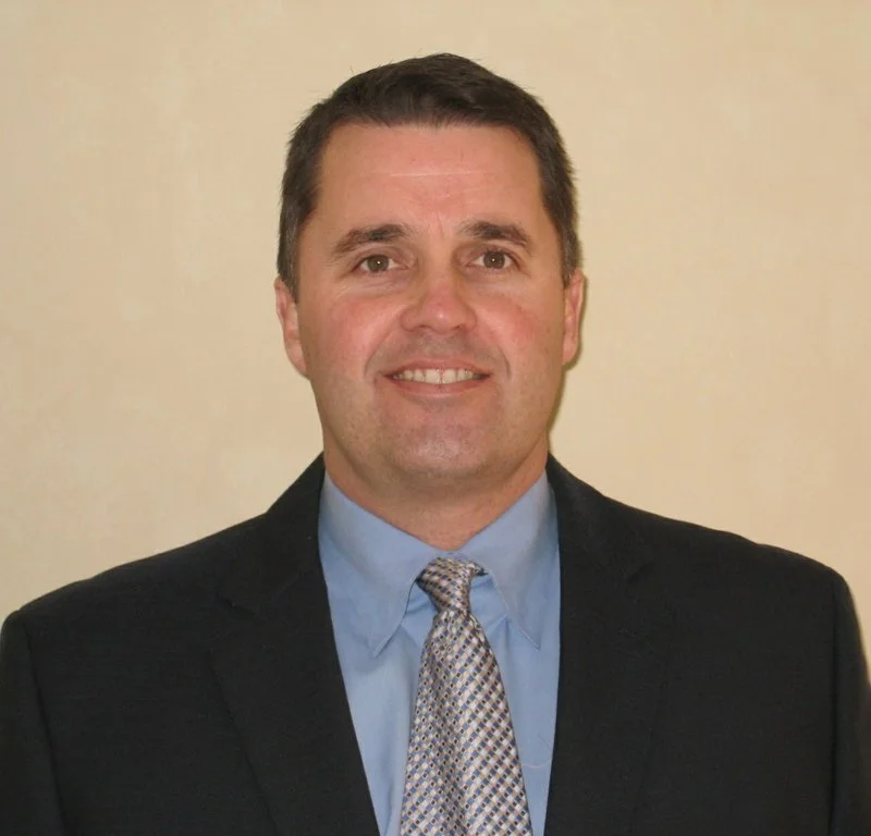 A man in a dark suit with a light blue shirt and patterned tie, smiling, against a plain beige background.