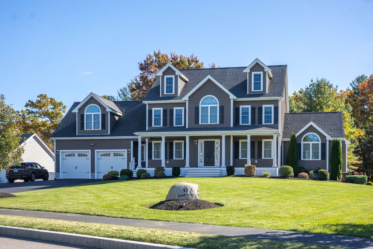 A large, two-story house with a gray exterior, white trim, and a front porch, surrounded by a neatly manicured lawn and trees in the background.