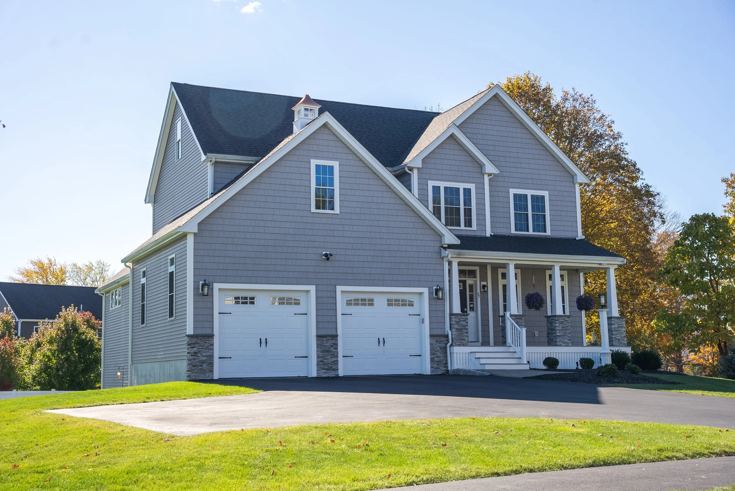 A two-story gray house with white trim, a front porch, and a driveway leading to two garage doors. The yard has green grass and trees in autumn colors.