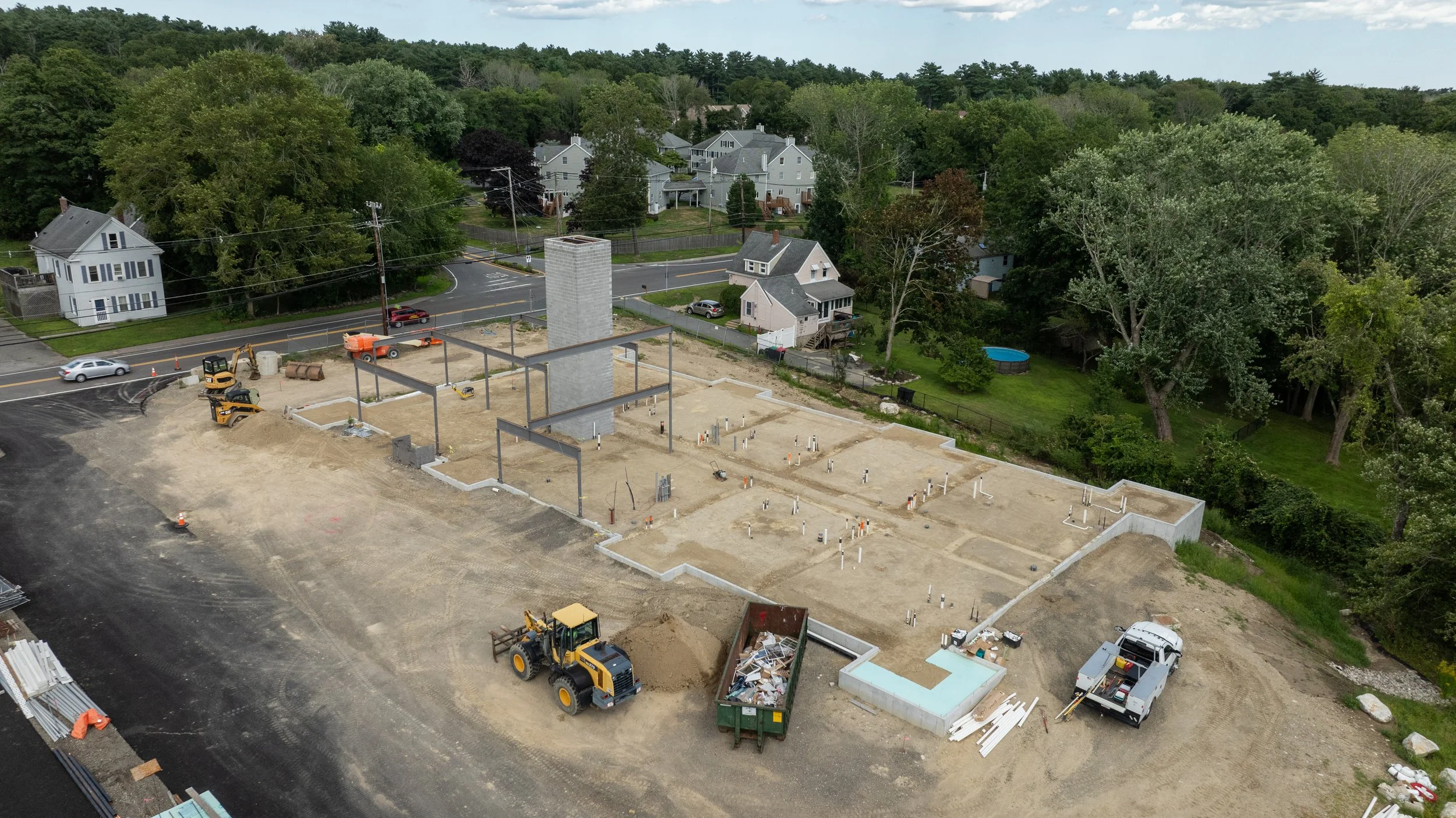 Construction site with foundation and steel beams, surrounded by trees and residential houses in the background.