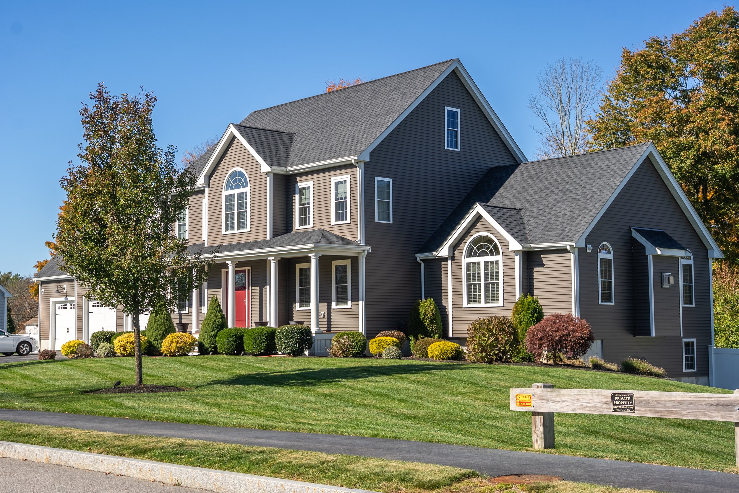 A large, modern, gray house with white trim, double garage, and front porch with red door, surrounded by a well-manicured lawn and colorful bushes, with trees and a blue sky in the background.