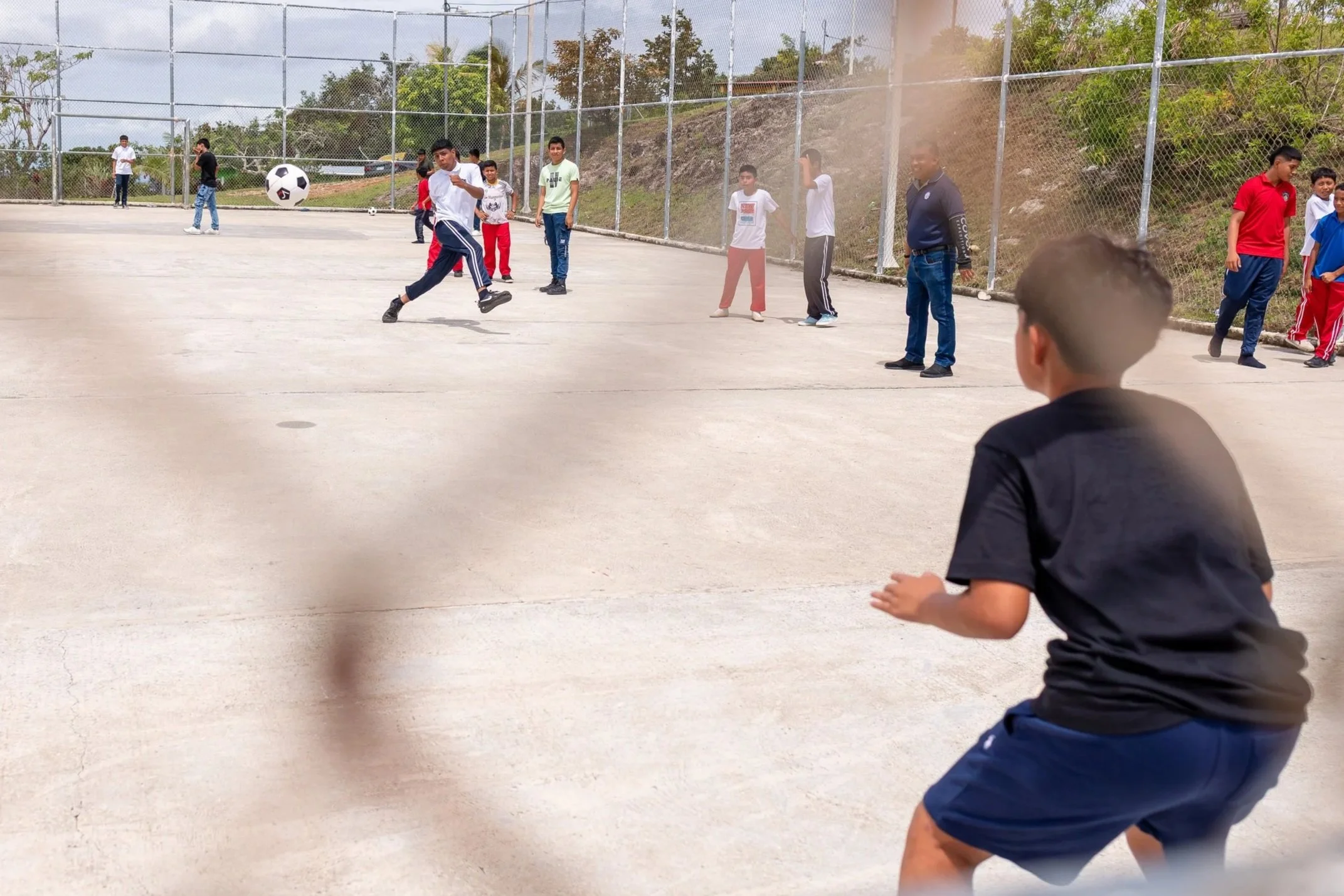 Niños jugando fútbol en un campo al aire libre, algunos observando y otros en acción, rodeados de un cerco de cadena y con árboles y cielo nublado en el fondo.