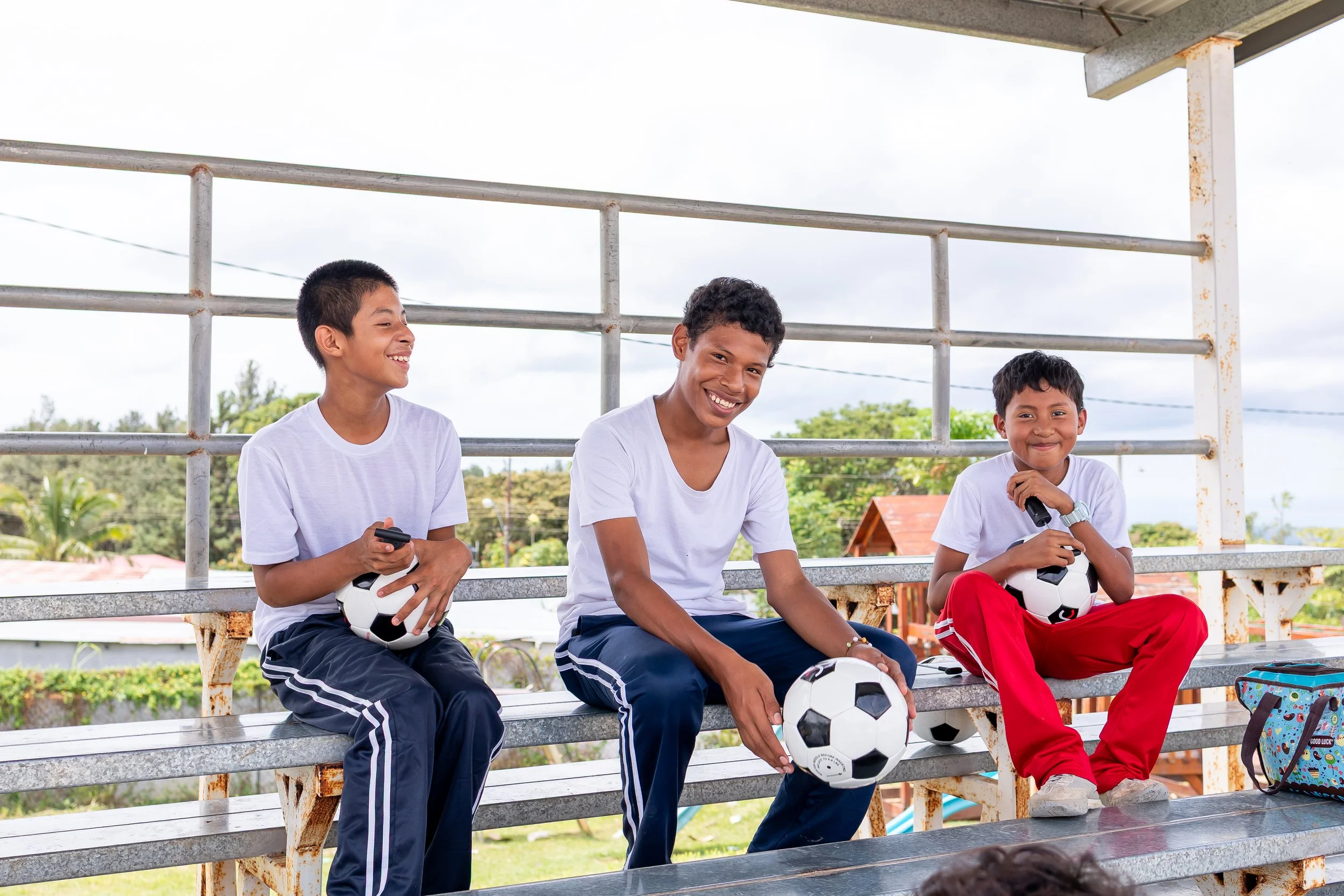 Tres niños sentados en las gradas de un estadio, sosteniendo balones de fútbol y microfonos, sonriendo y vistiendo ropa deportiva blanca, azul y roja.