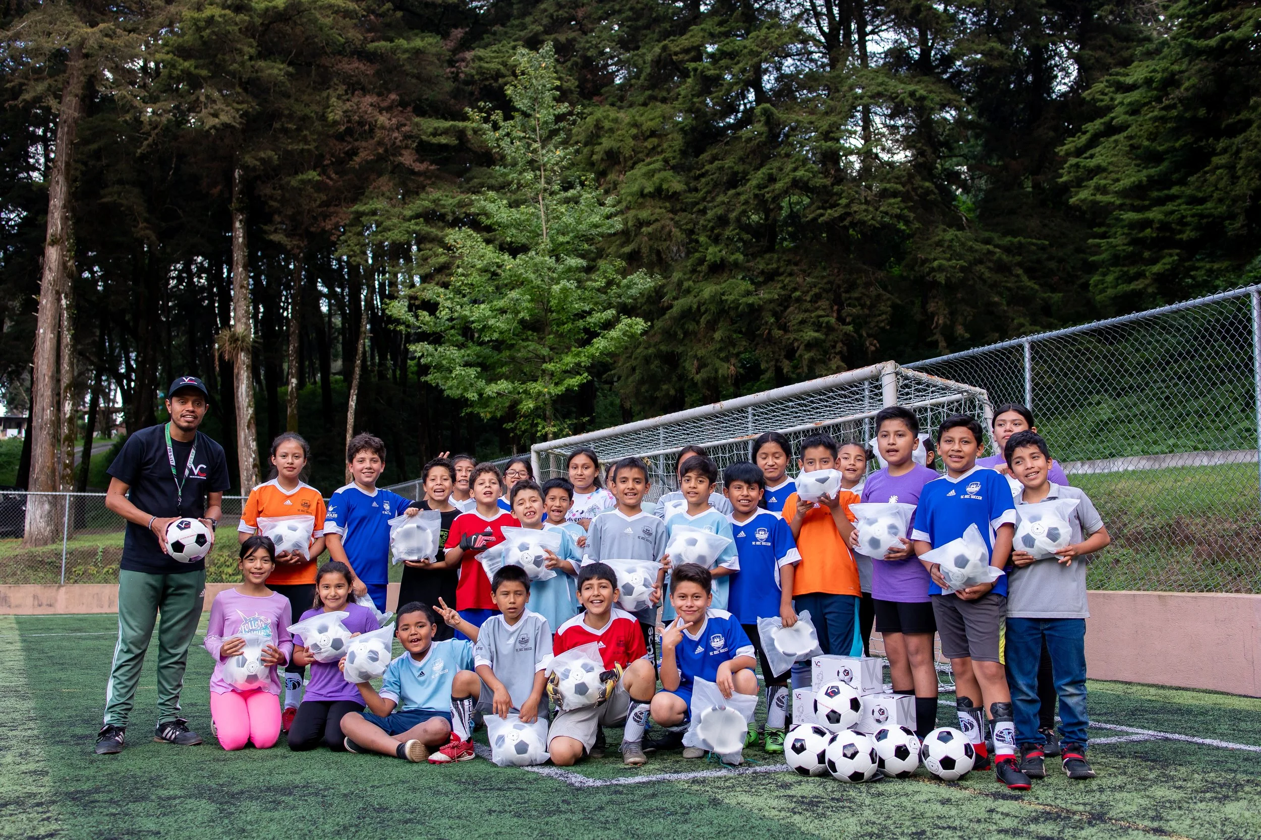 Grupo de niños y una persona adulta en un campo de fútbol, con algunos niños sosteniendo balones y paquetes, parece un evento escolar o de equipo juvenil de fútbol, en un entorno de parque con árboles en el fondo.