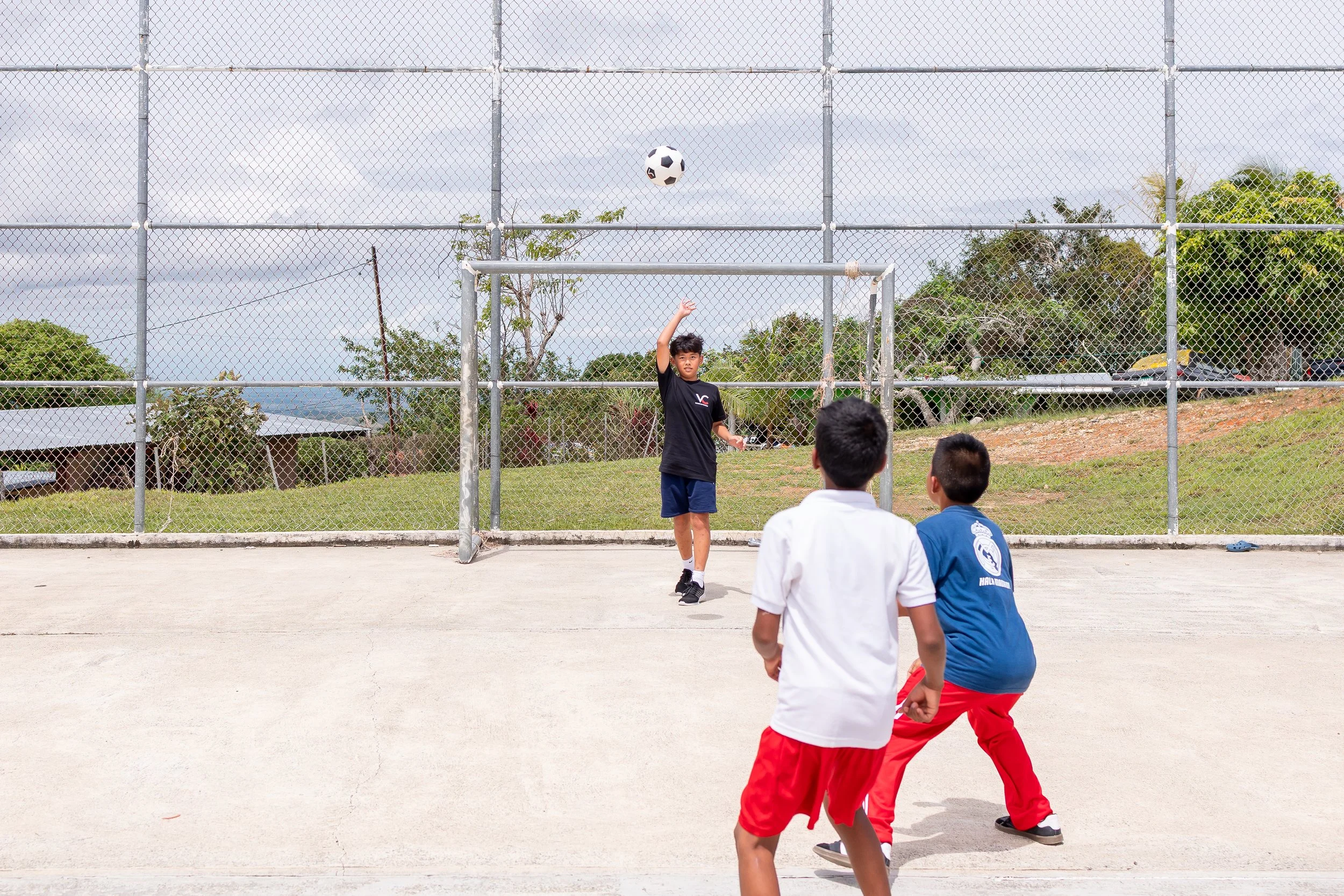 Tres niños jugando a la pelota en un área al aire libre con cercas y árboles en el fondo.
