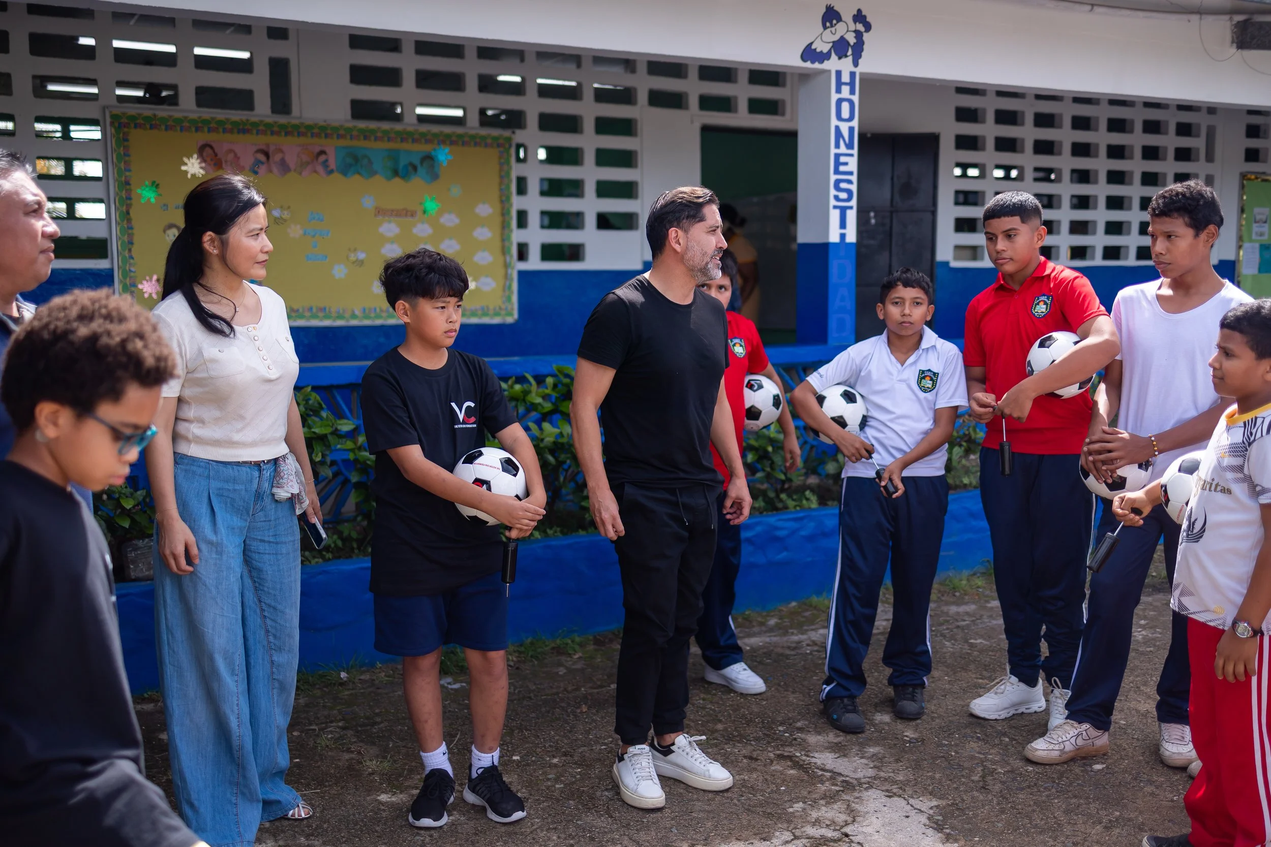 Personas adultas y niños en un círculo en una cancha de tierra, algunos con balones de fútbol, discutiendo o aprendiendo en un entorno escolar, con un cartel de fondo que dice 'Honestidad' y decoraciones en la pared.