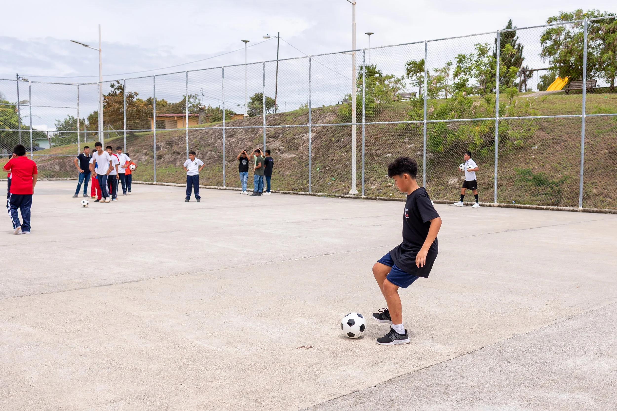 Niños jugando fútbol en una cancha al aire libre, algunos con balones y otros en grupos, en un día nublado.