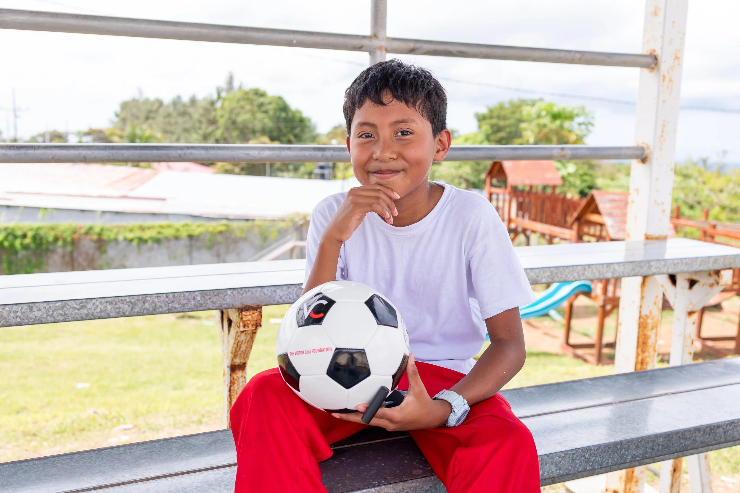 Niño sonriendo sentado en banca, sosteniendo balón de fútbol en un ambiente al aire libre.