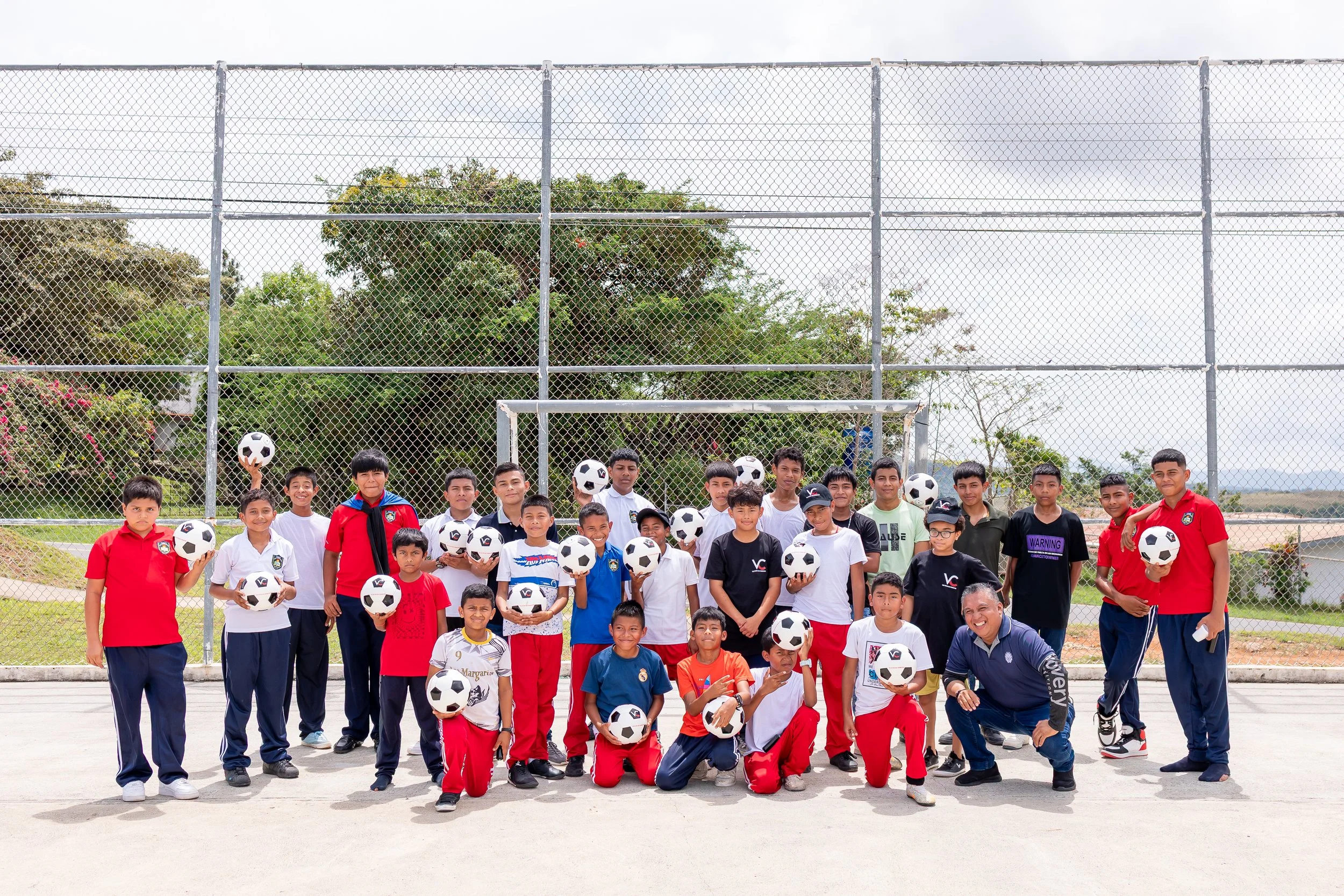 Grupo de niños y adultos en un campo de fútbol, algunos sosteniendo balones, en un día soleado con fondo de árboles y una cerca de malla metálica.