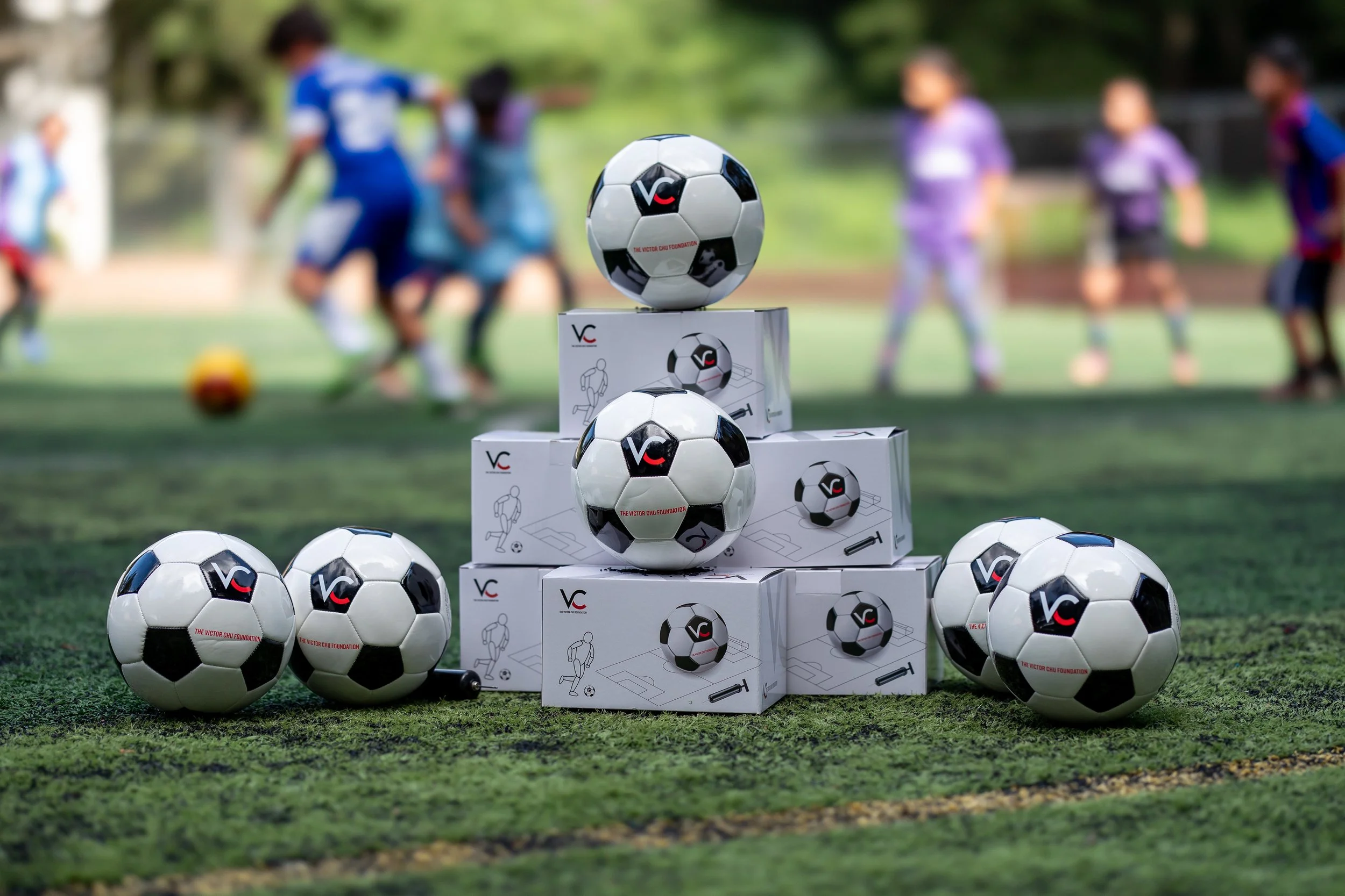 Balones de fútbol y cajas en un campo, con niños jugando en el fondo.