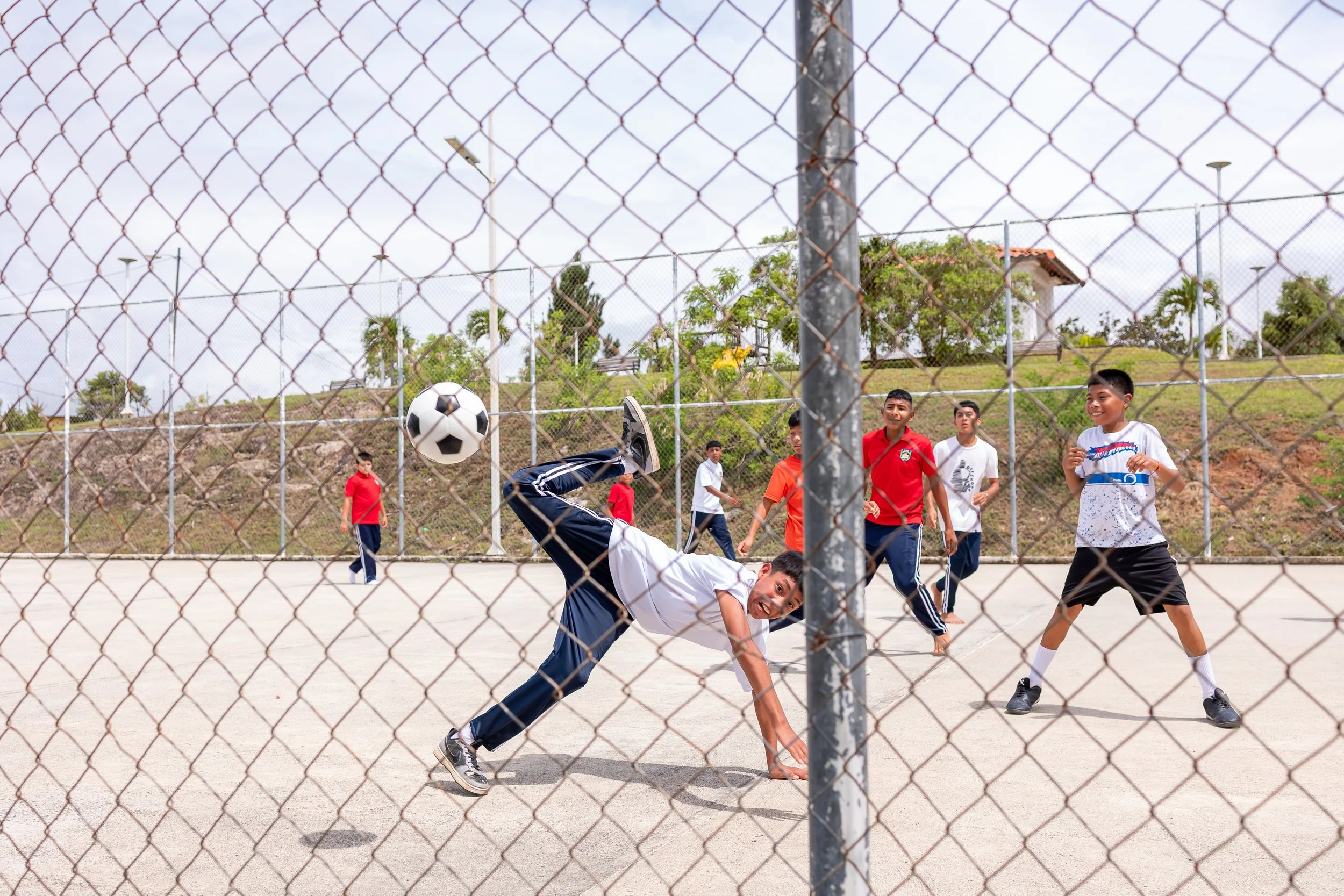 Niños jugando fútbol en una cancha al aire libre, algunos en movimiento y uno en el suelo, detrás de una cerca metálica.