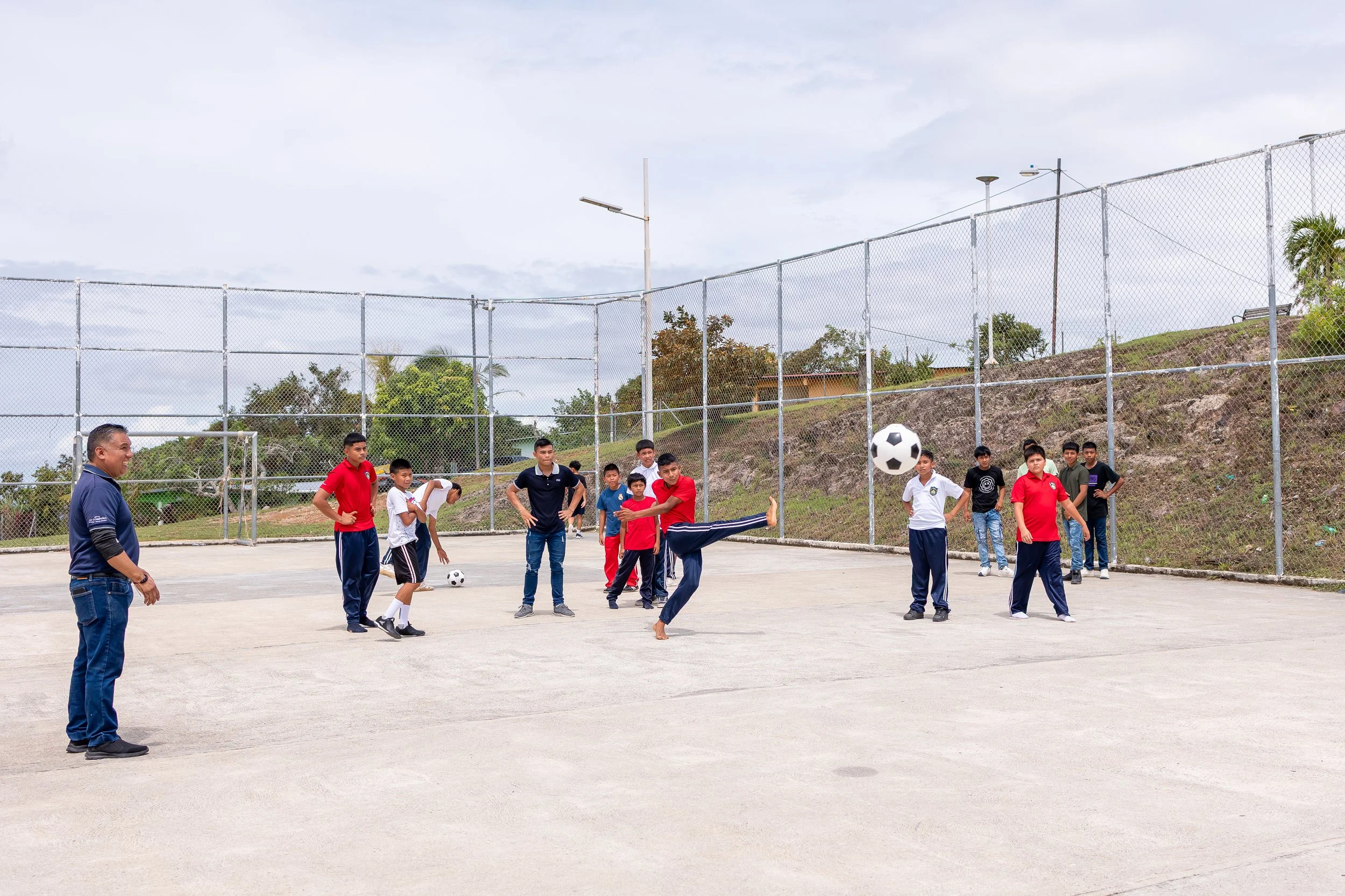 Grupo de niños y un adulto en un recreo jugando fútbol en una cancha rodeada de mallas, con un cielo nublado y naturaleza en el fondo.