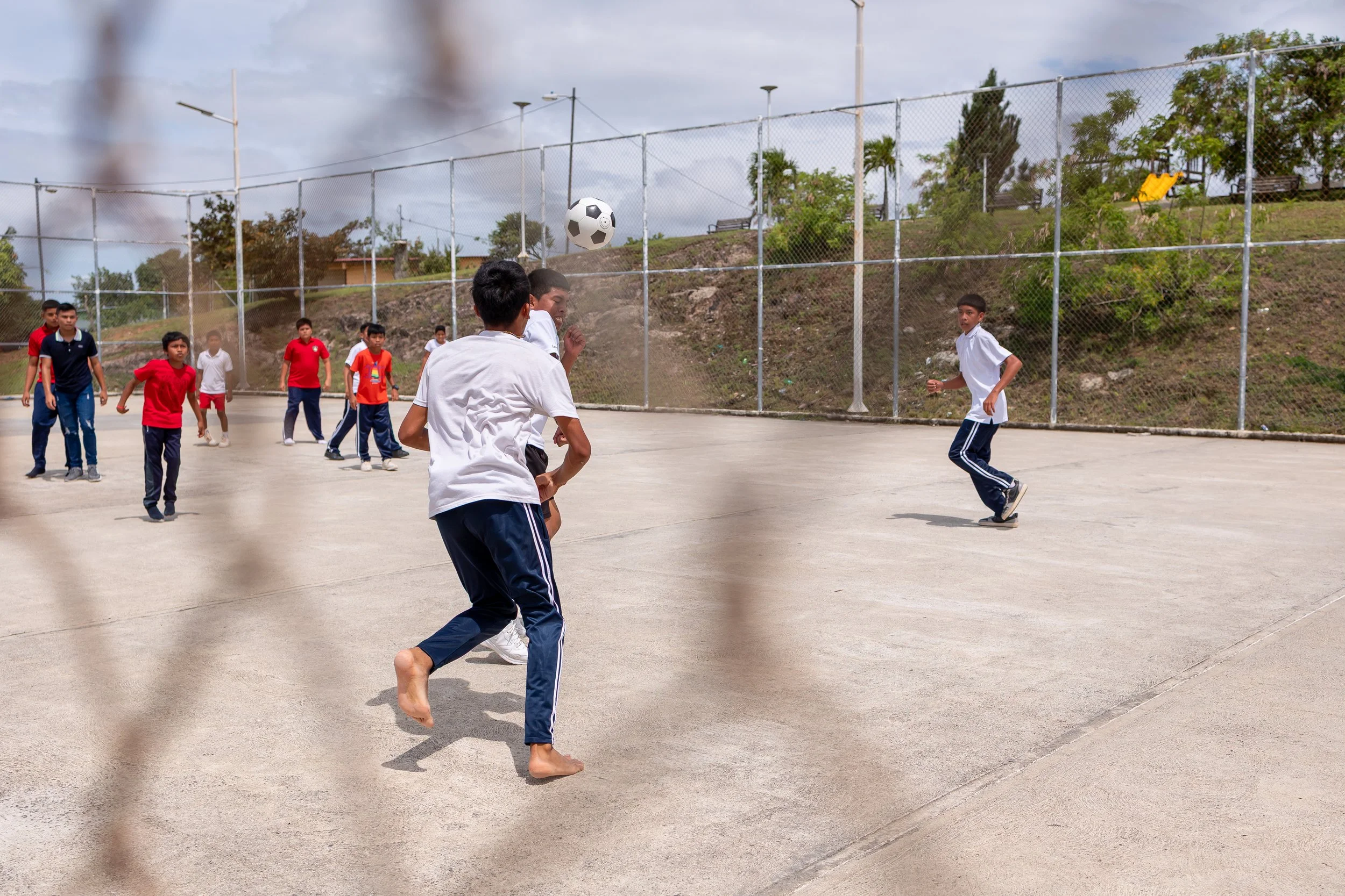 Niños jugando fútbol en una cancha al aire libre, algunos en fila y otros en movimiento, con árboles y una cerca de malla al fondo.