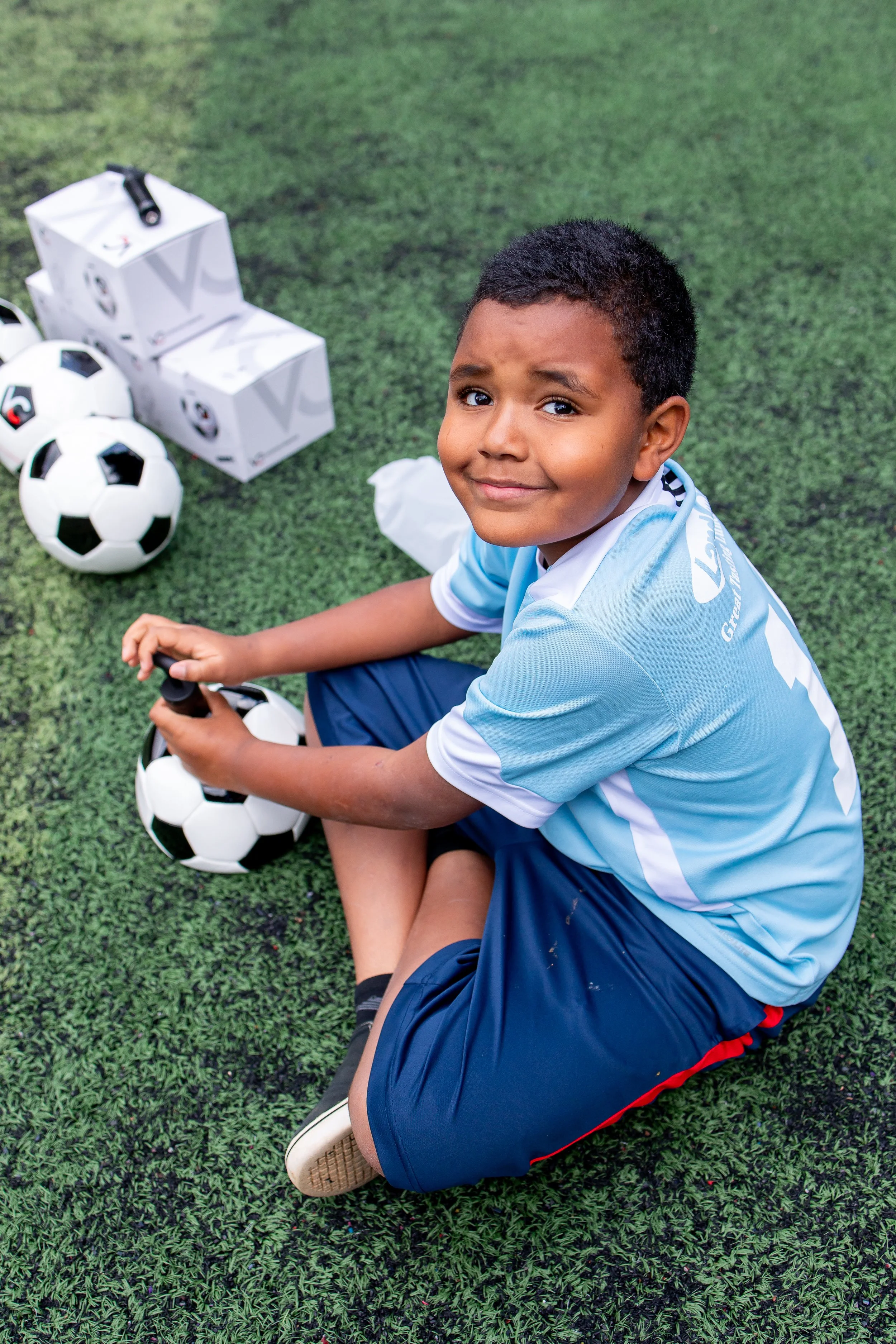 Niño en uniforme de fútbol sentado en césped con balones y cajas de jugo.
