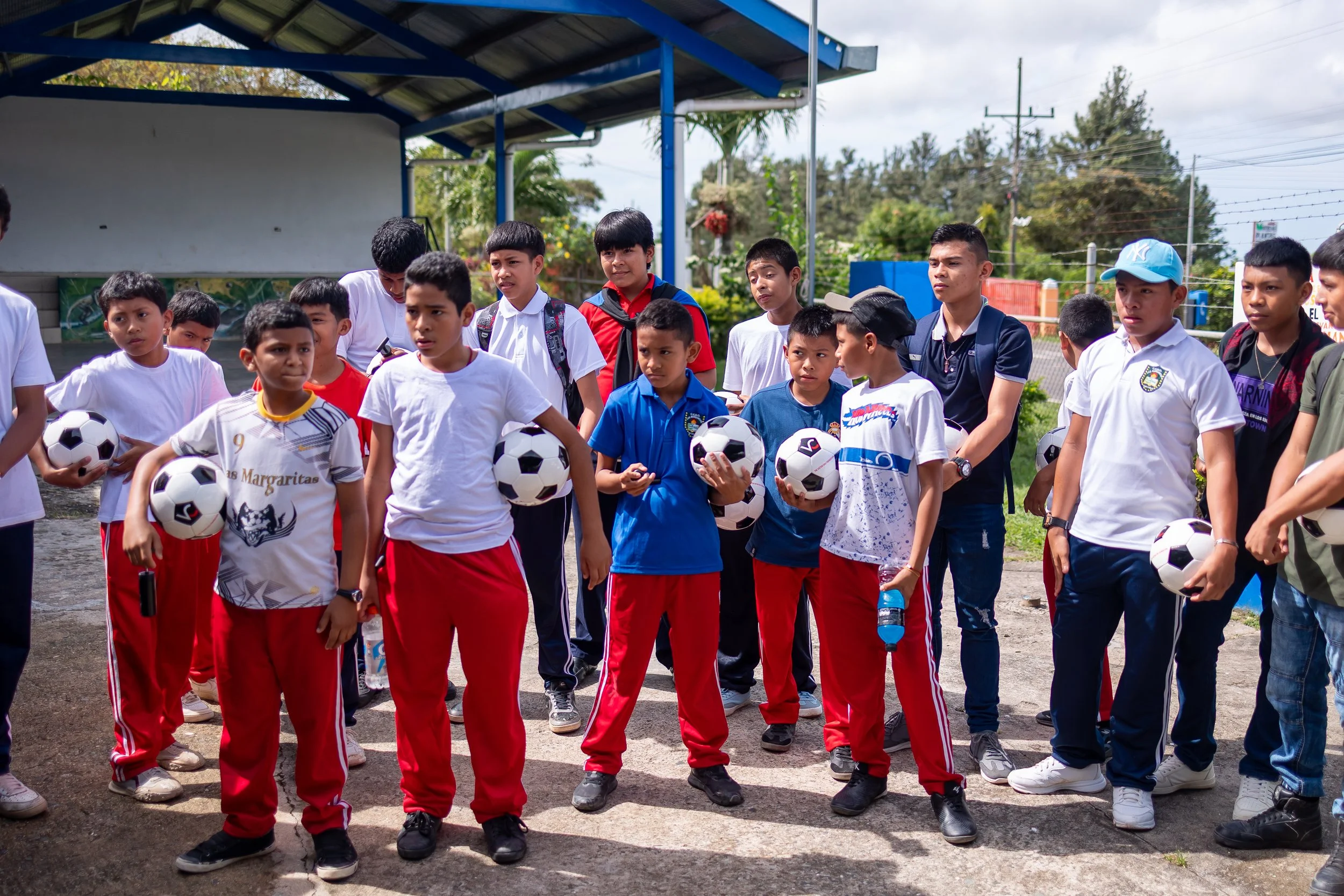 Grupo de niños en una cancha de fútbol, algunos sosteniendo balones, en una especie de reunión o entrenamiento al aire libre bajo un toldo azul.