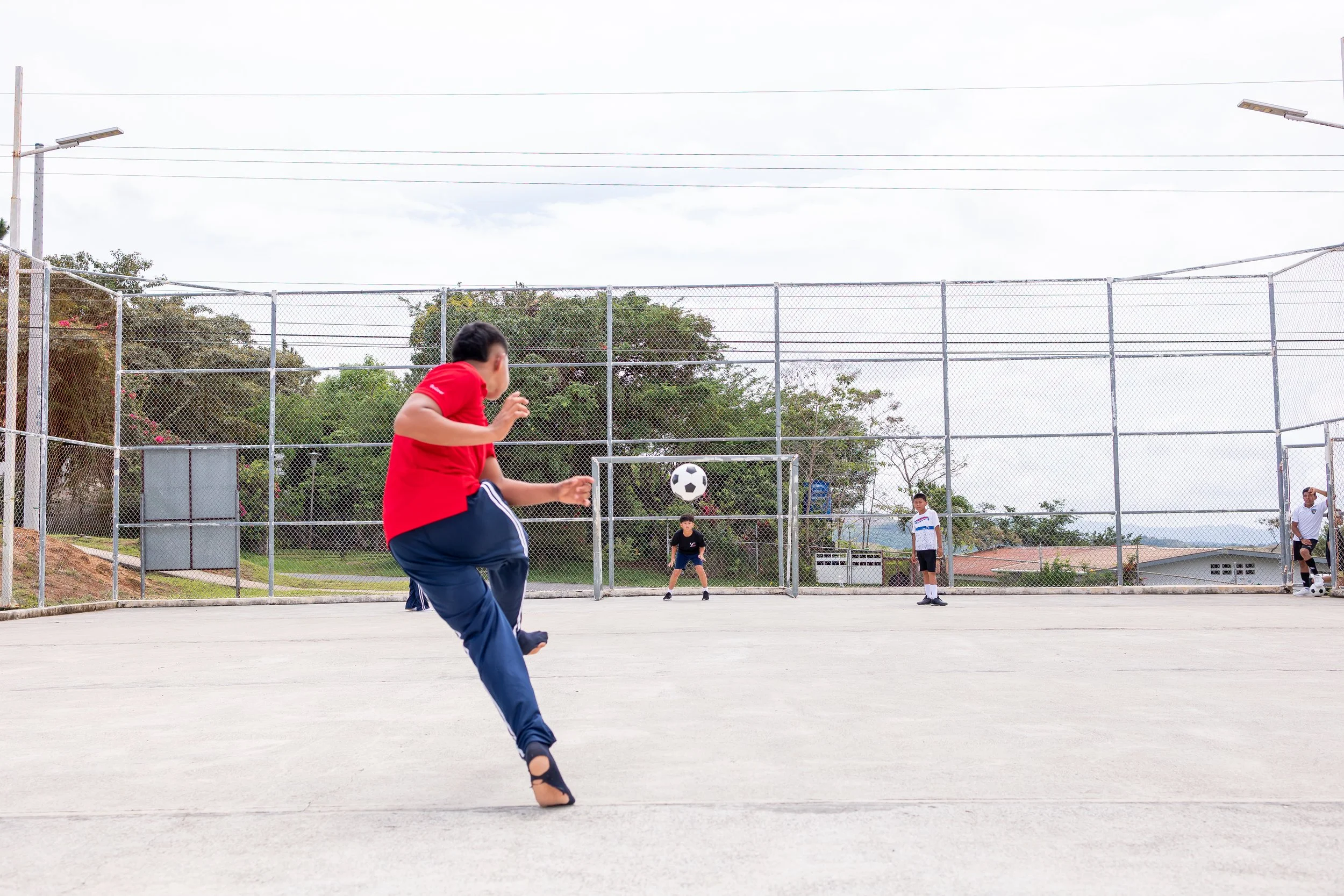Niño en camiseta roja y pantalones azules jugando fútbol en una cancha exterior, con al fondo otros niños y árboles.