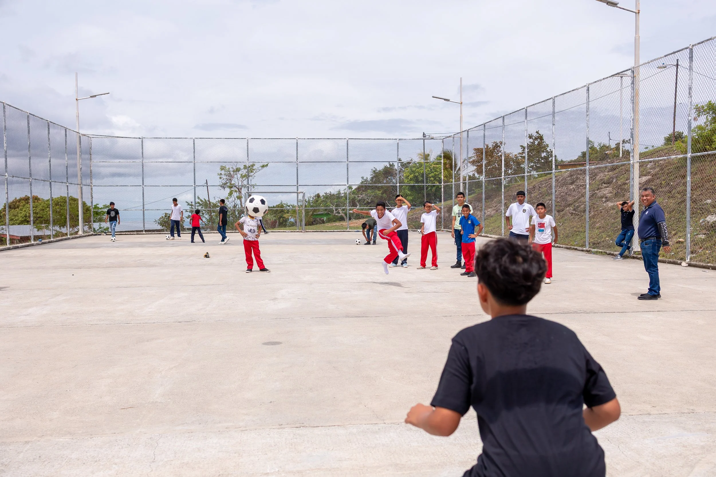 Niños y adultos jugando fútbol en un campo abierto con malla de alambre y rodeados de naturaleza.