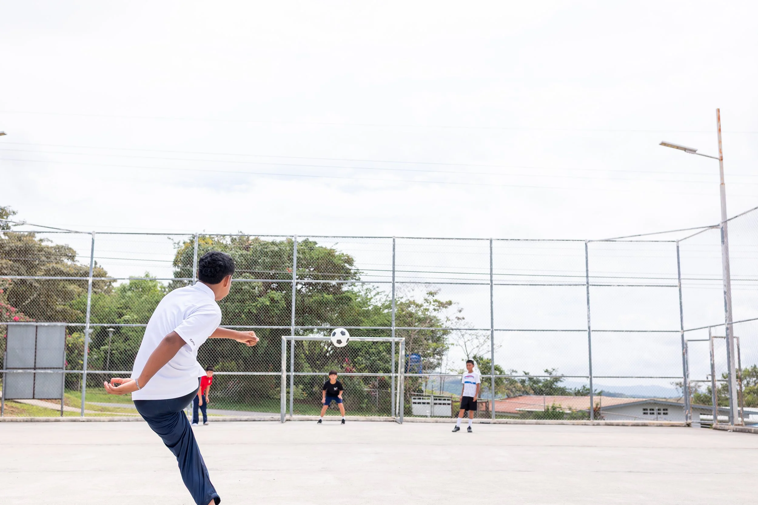 Niños jugando fútbol en una cancha exterior, uno de ellos está a punto de patear el balón.