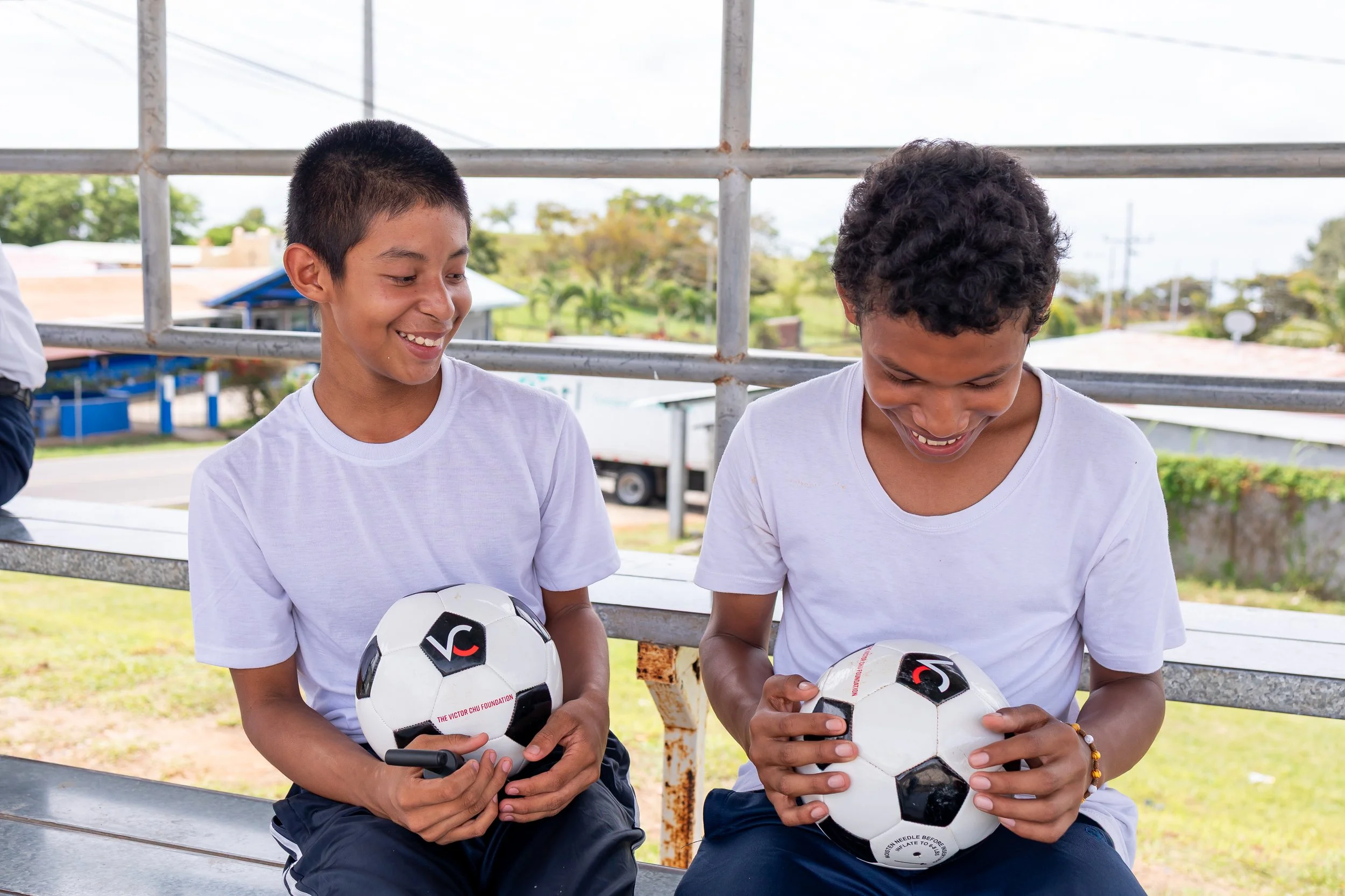 Dos niños sonrientes, con camisetas blancas, sentados en un banquillo y sosteniendo balones de fútbol.