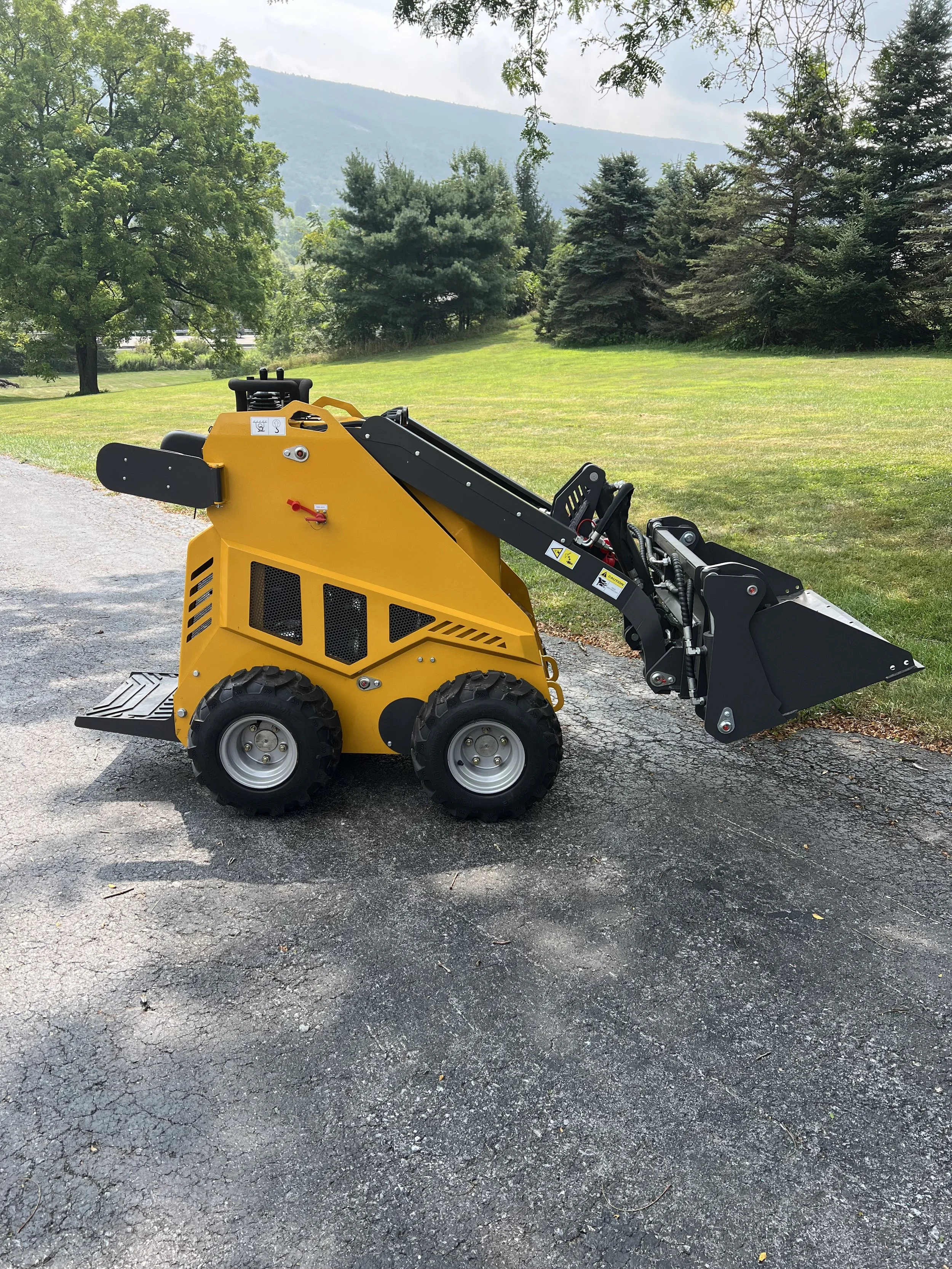 A small yellow industrial skid steer loader with a black attachment, parked on a paved surface beside a grassy area with trees and mountains in the background.