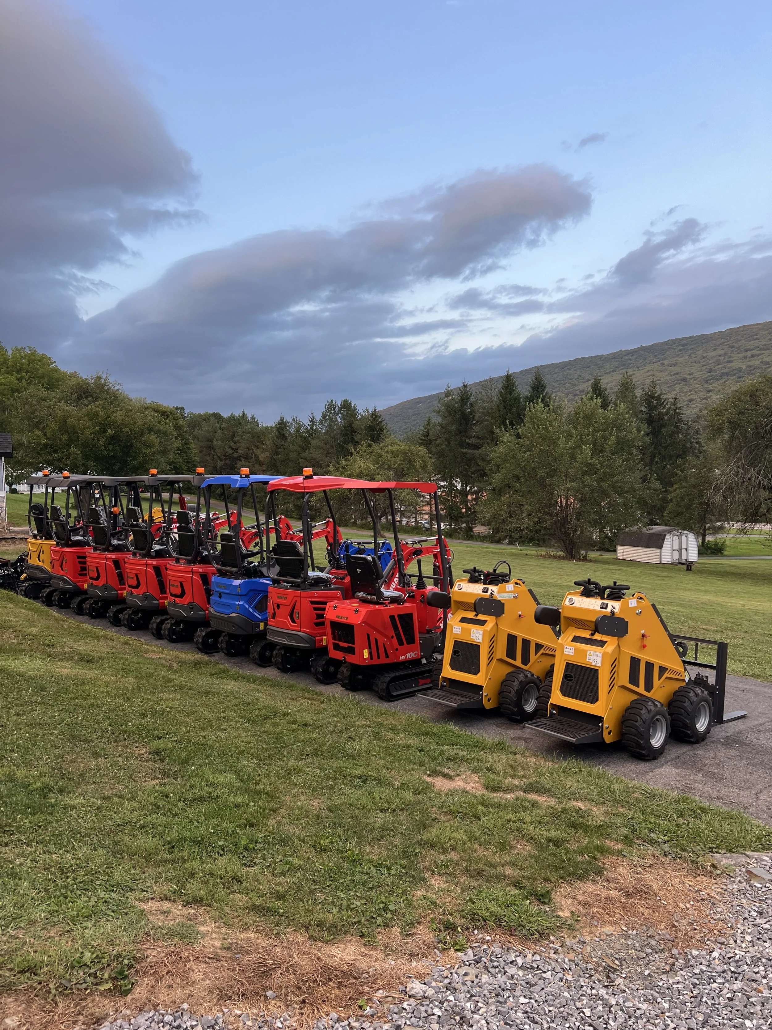 Line of small construction vehicles, including red, blue, and yellow machinery, parked on a gravel path in a grassy area with trees and mountains in the background.