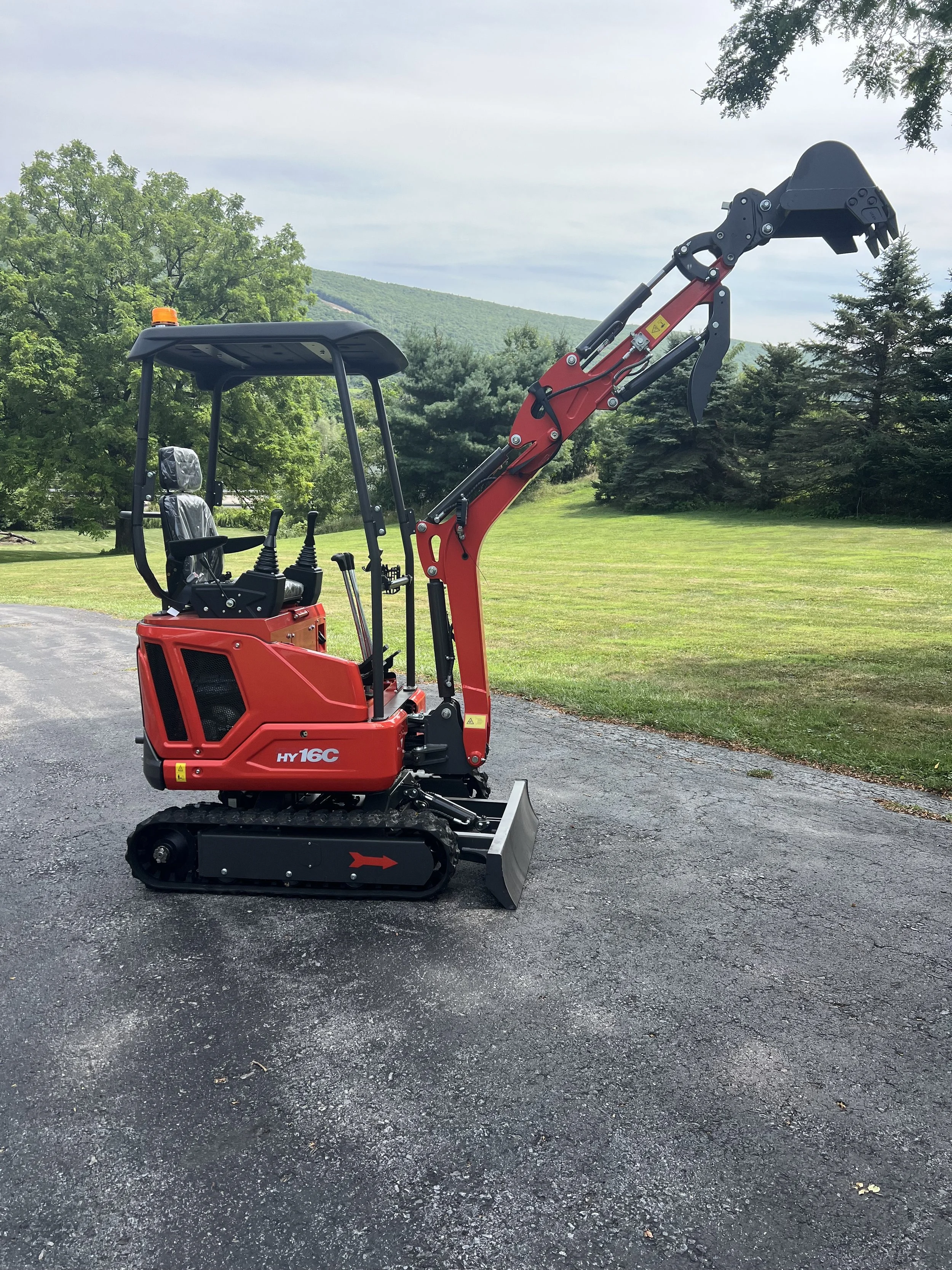 Red mini excavator with tracked wheels parked on asphalt surface in a green outdoor setting.