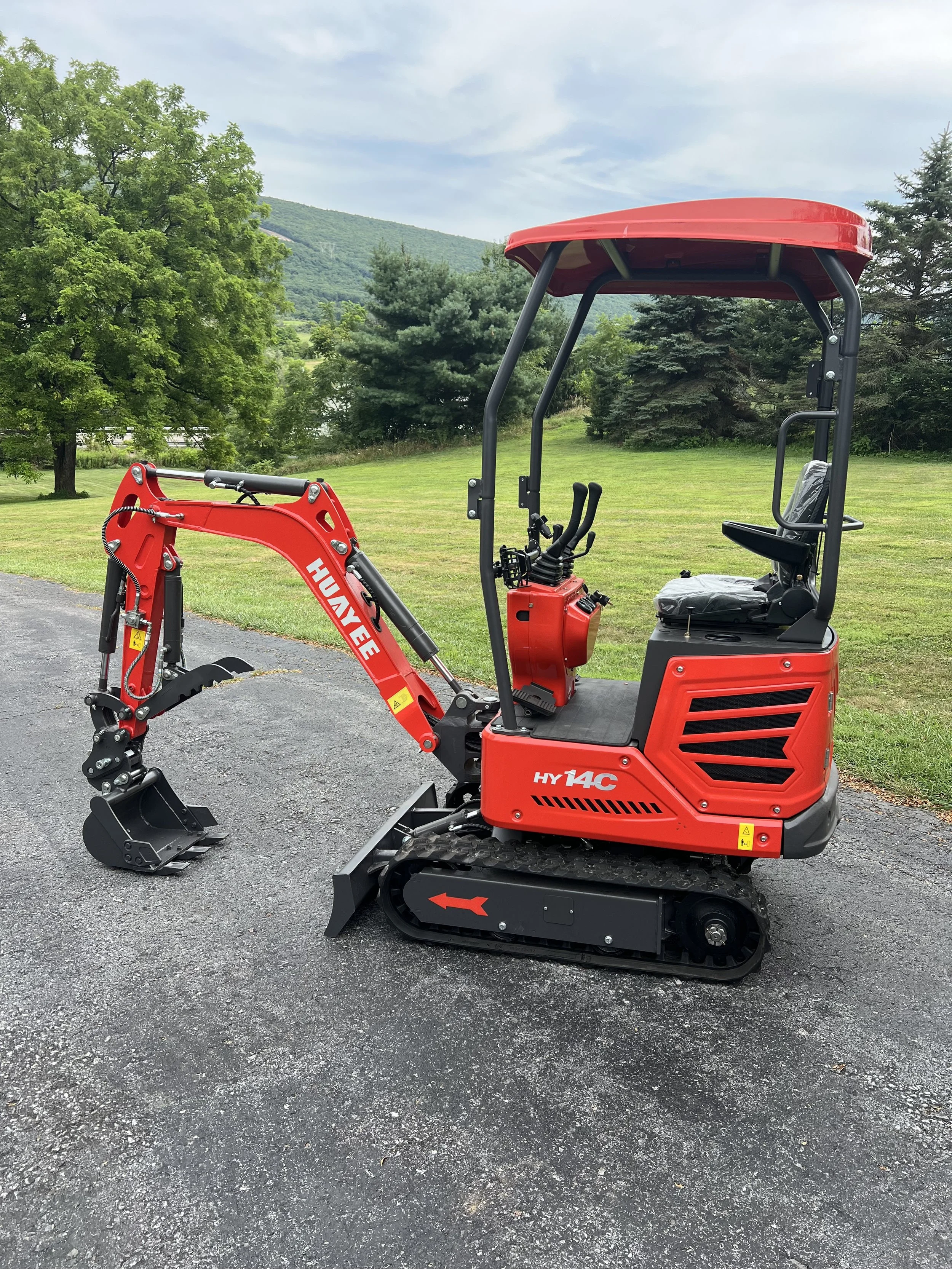Red compact excavator with black tracks parked on gravel in a lush green outdoor setting.