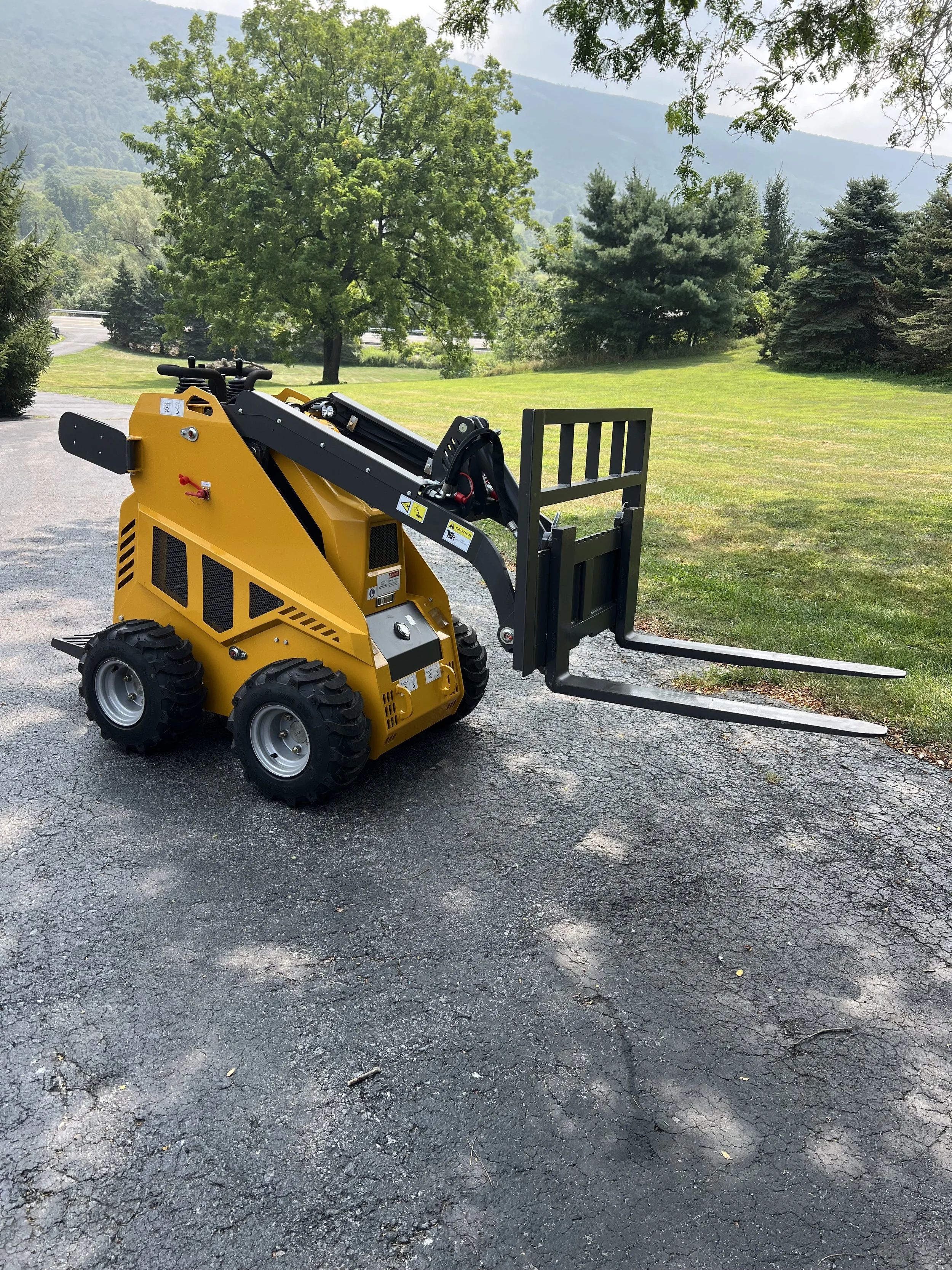 Yellow mini skid steer loader with forks attachment on a paved driveway, surrounded by trees and greenery with mountains in the background.