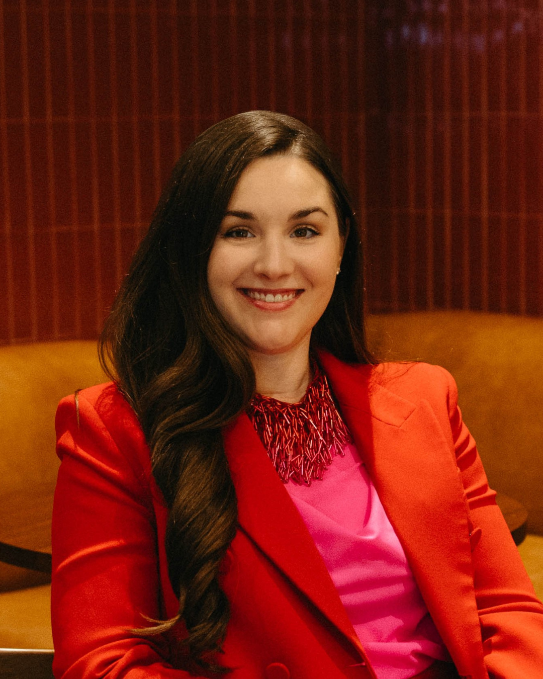 Ashlyn Holeyfield, Parapet Advisors. A woman with long dark hair smiling, wearing a red blazer and a pink top, sitting in a room with a red and brown background.