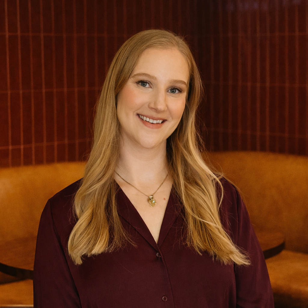 Emily Gershon, Parapet Advisors. A young woman with long blonde hair smiling, wearing a burgundy blouse and a necklace, indoors with a red patterned background and a yellowish-brown sofa.