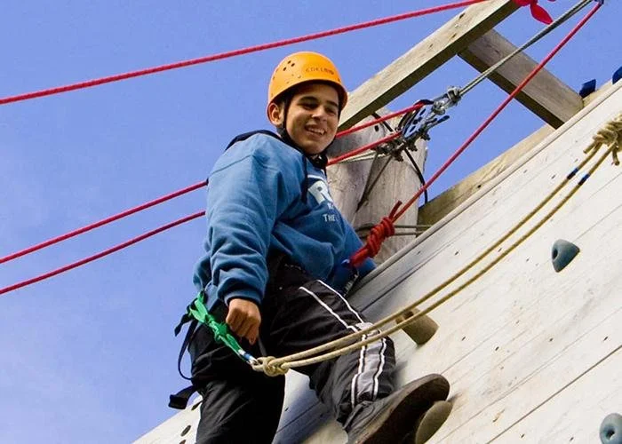 Young boy in a blue jacket and orange helmet climbing an outdoor rock wall with red and yellow ropes, smiling.