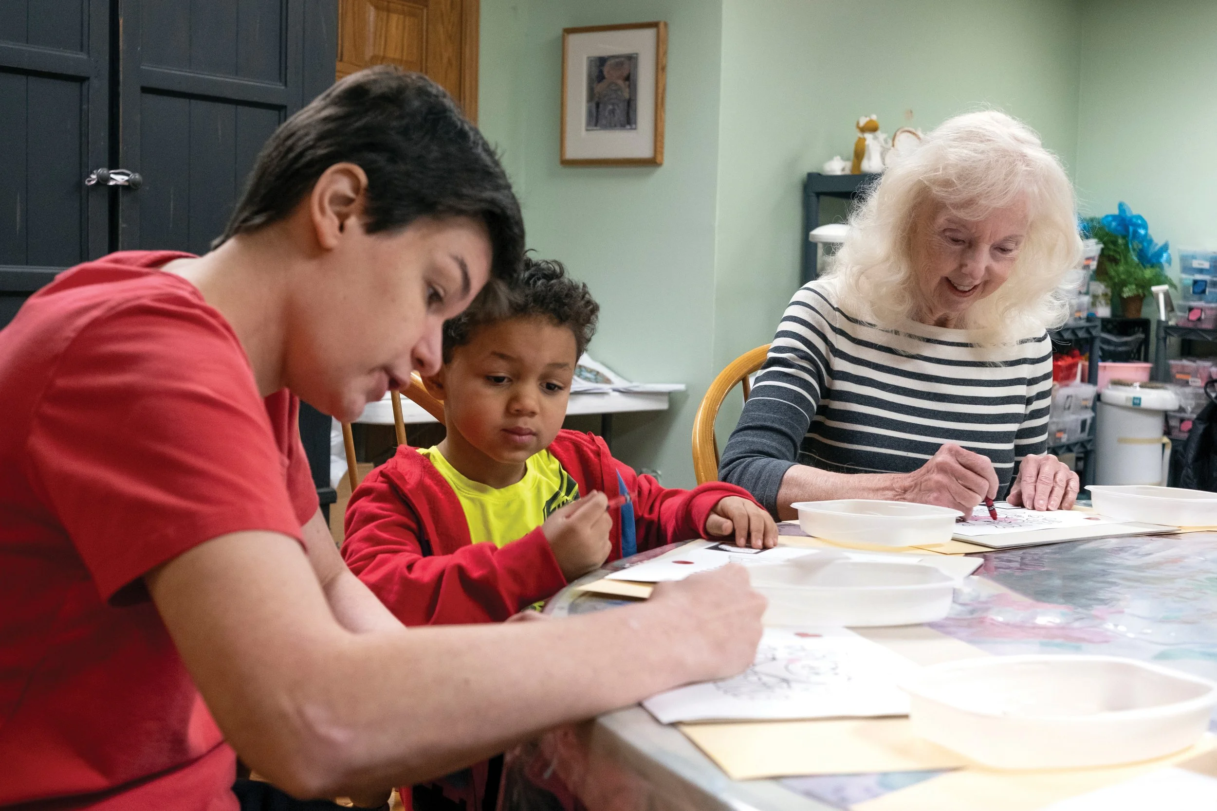 Three people, a young man, a young boy, and an elderly woman, sit at a table engaged in arts and crafts. The elderly woman is smiling while working on her project, and the children are focused on their activities.