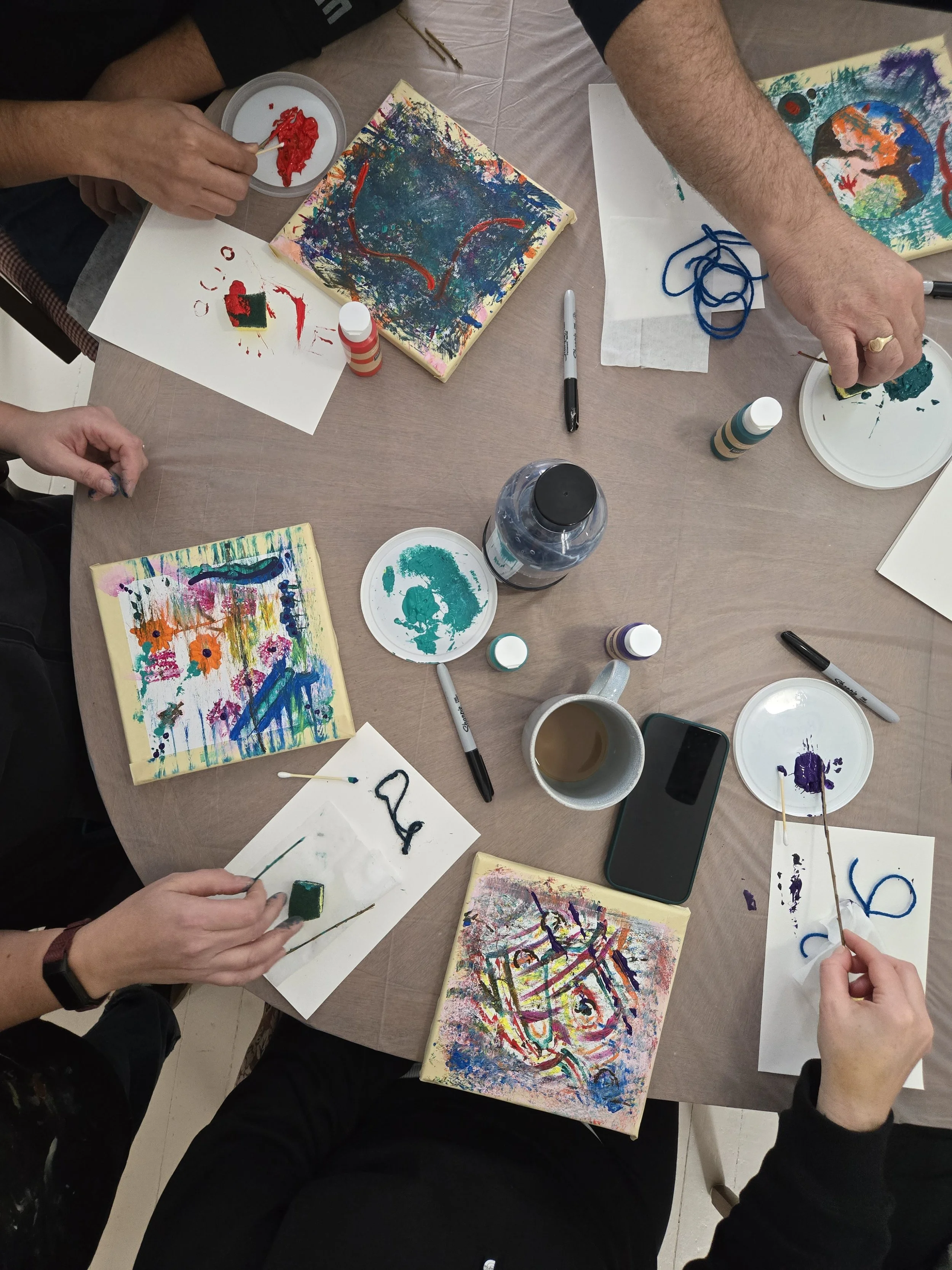 People painting colorful abstract art on small canvases at a round table with paints, brushes, markers, and cups of coffee.