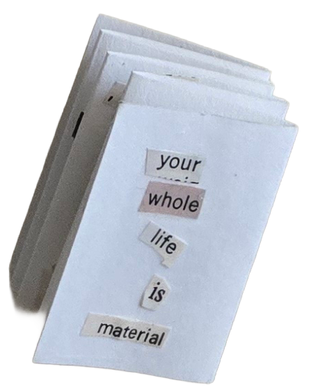 A folded paper book with cut-out words that spell 'your whole life is material' arranged vertically.
