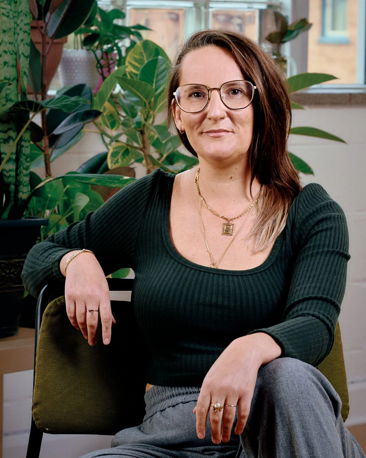 A woman with brown hair, wearing glasses, a black ribbed sweater, and jewelry, sitting in a chair indoors with green plants and a window in the background.