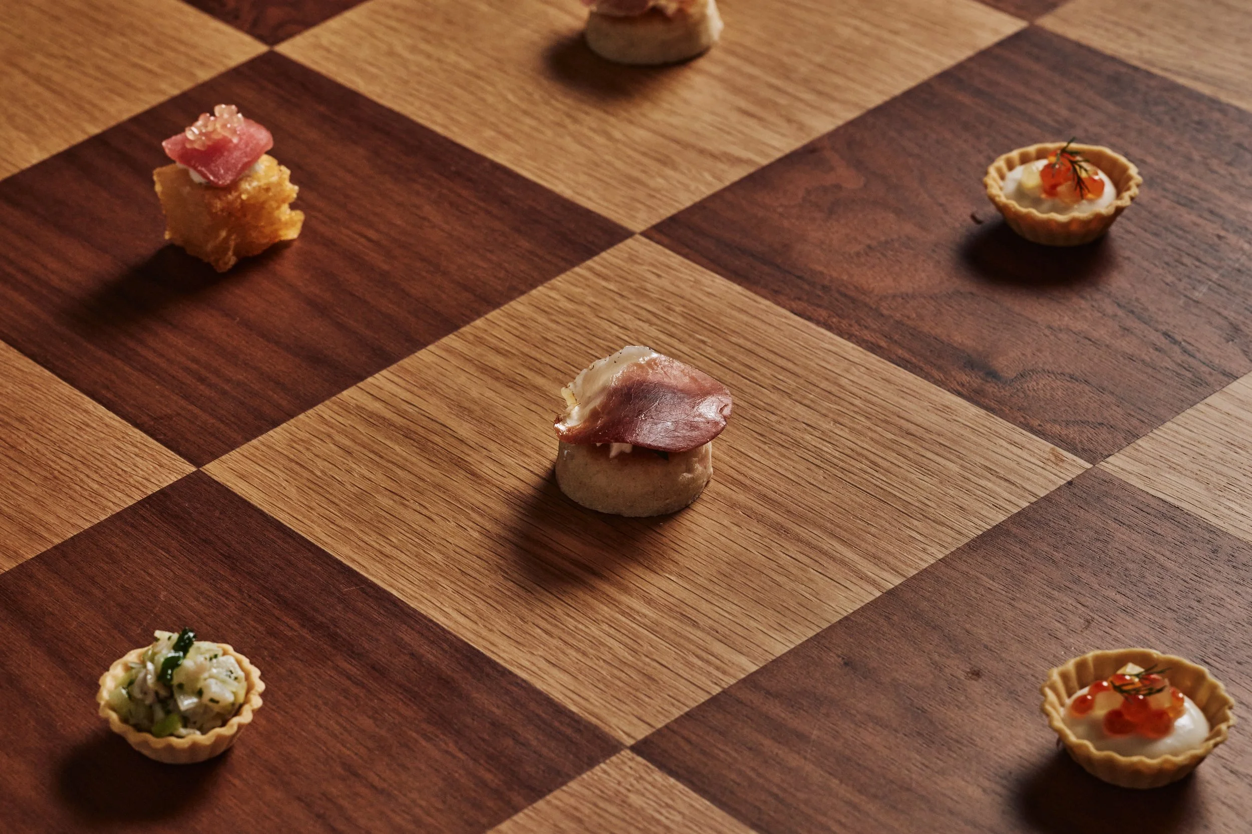 Various small gourmet appetizers on a wooden checkered serving board, including tartlets and pieces of cured meats.