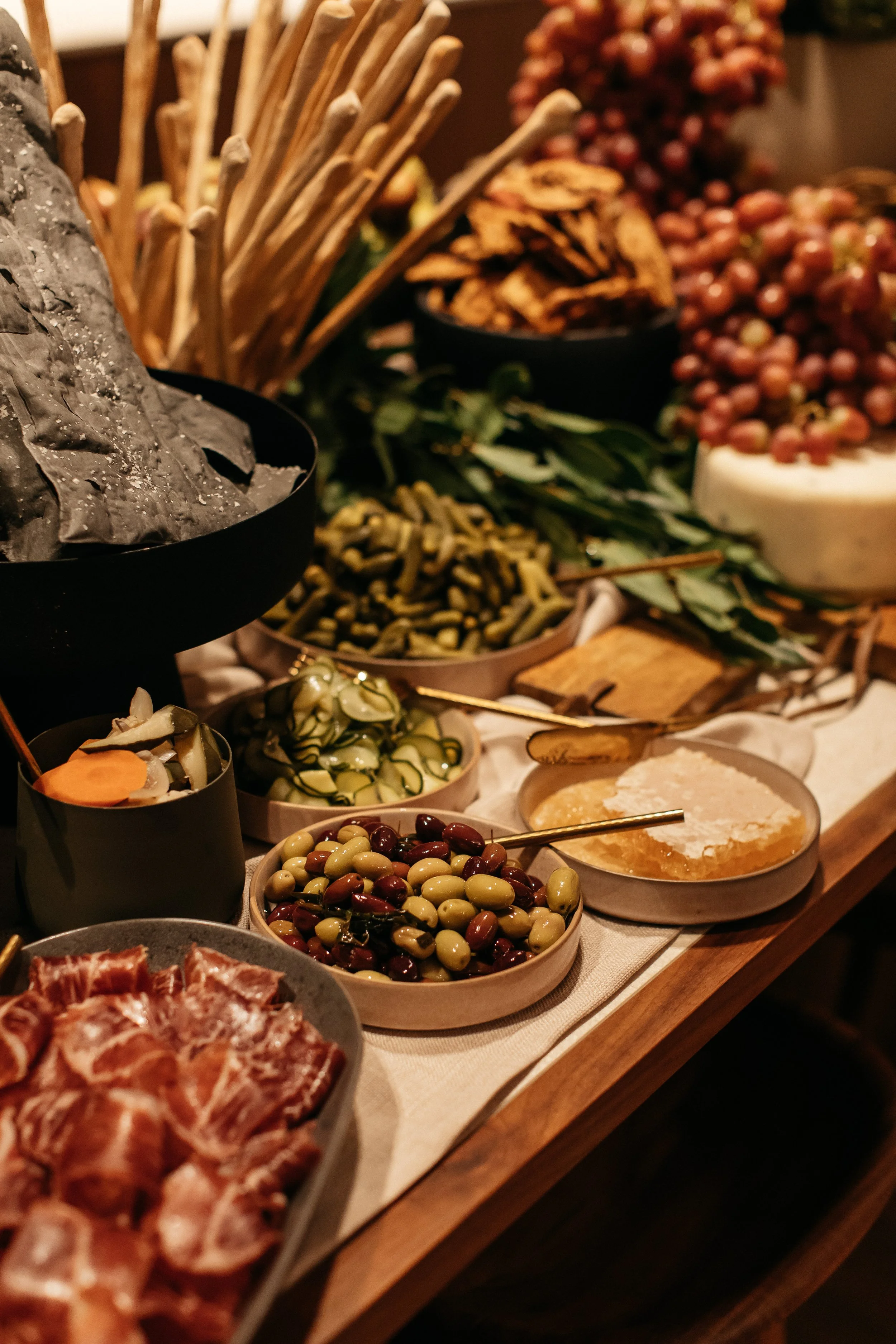 Assorted bowls of olives, cured meats, cheeses, and breadsticks on a wooden table, likely part of a charcuterie or antipasto platter.